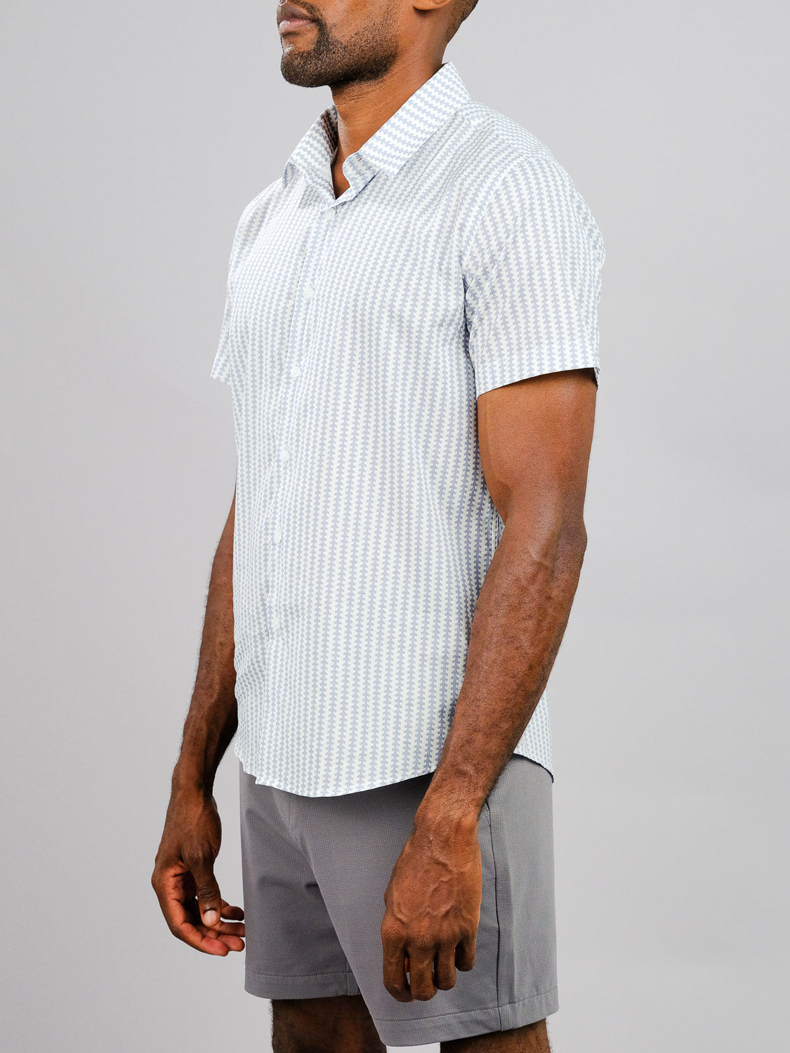 A man wearing the Shima Printed Cotton Shirt by ID Menswear and gray shorts stands against a plain light gray background, his head slightly turned and hands relaxed at his sides.