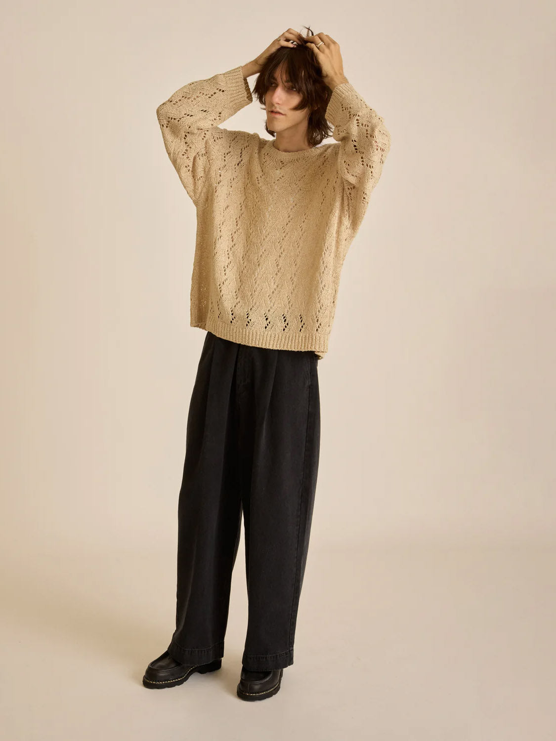 Against a plain background, a person dons the loose Obey Shimoda Sweater in beige with wide-leg dark pants. Their shaggy hair and hands on their head add to their casual vibe, completed by black shoes.