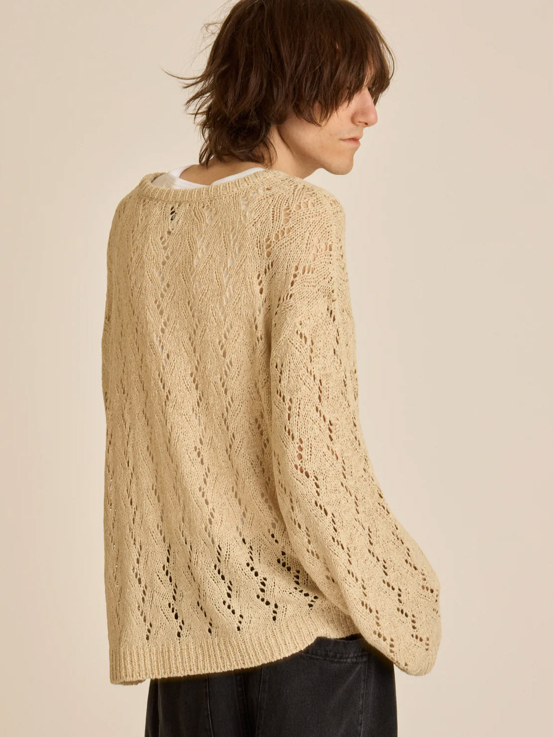 A person with shoulder-length brown hair wears the Obey Shimoda Sweater, a cream-colored, relaxed knit piece over a white shirt. They face slightly sideways with a neutral expression, paired with dark pants. The plain beige background enhances the outfits cozy charm.