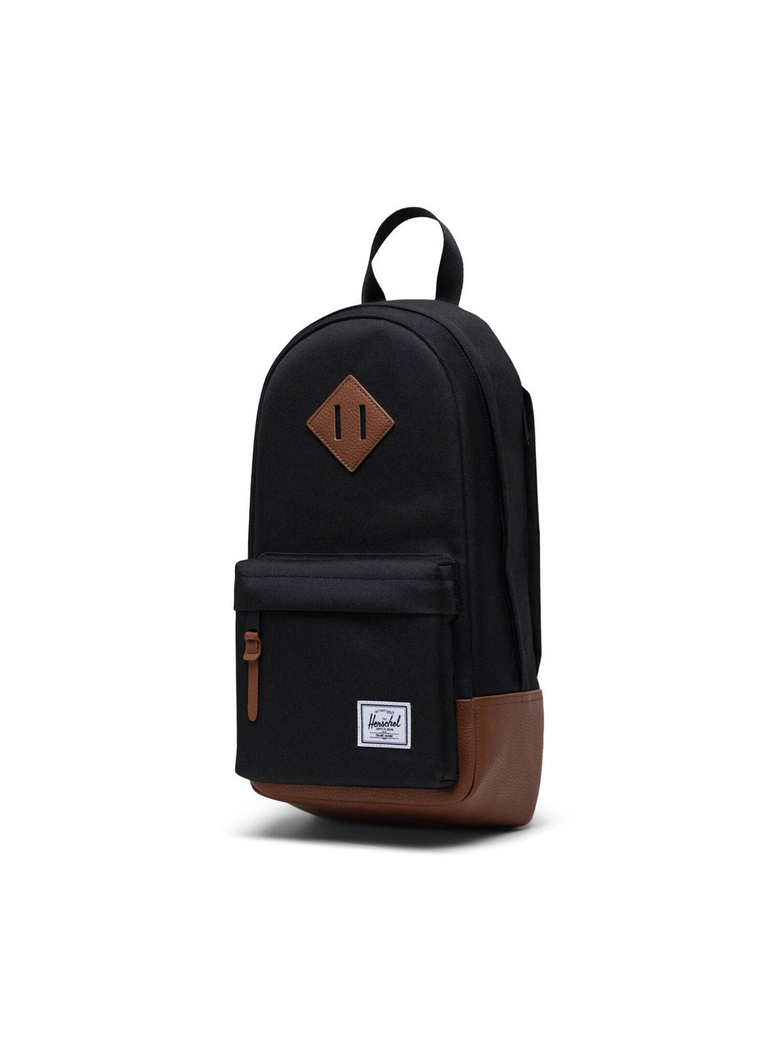 The Herschel Heritage Shoulder Bag in black features brown accents on the base and zipper pull, a front zip pocket, top handle, and a white rectangular logo patch on the front pocket, shown against a white background.