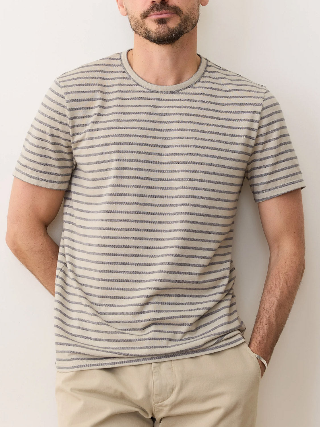 A bearded man wears Marine Layer’s Signature Jacquard Crew Tee with thin gray stripes and light tan pants, standing against a light-colored wall.
