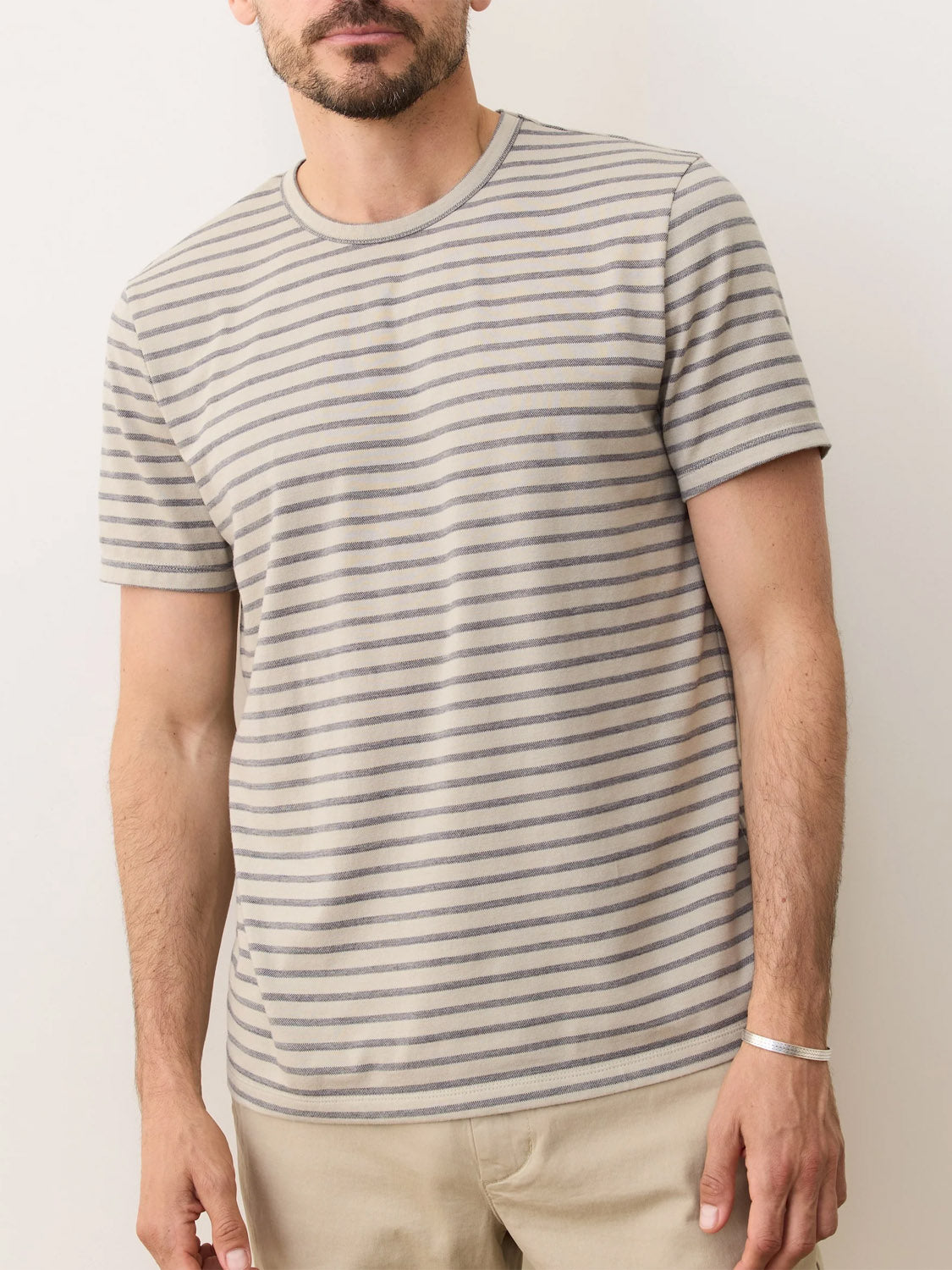 A man stands against a plain background wearing Marine Layer’s Signature Jacquard Crew Tee in light gray and beige stripes with light khaki pants. His face is partly out of frame, and he accessorizes with a thin silver bracelet.