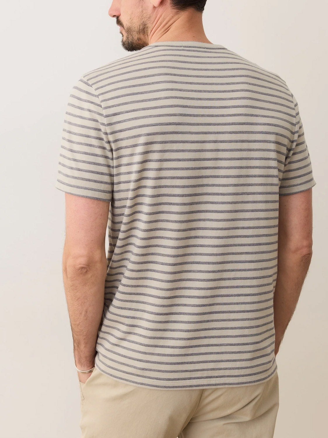 A man with short dark hair and a beard is seen from the back, wearing the Marine Layer Signature Jacquard Crew Tee and beige pants, standing against a plain light background.