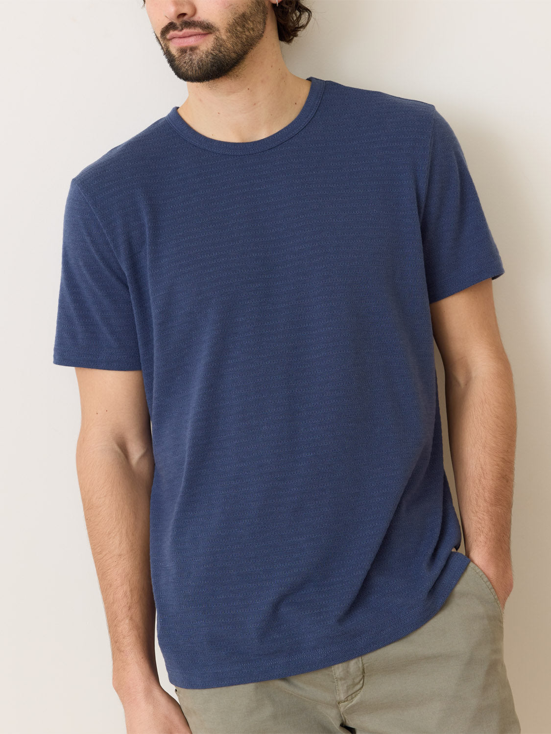 A bearded person in a Marine Layer Signature Jacquard Crew Tee and light pants stands against a neutral backdrop, one hand in a pocket, gazing slightly sideways.
