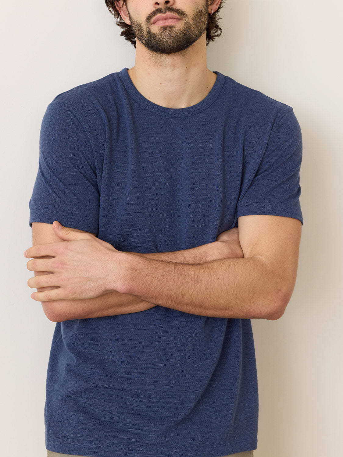 A bearded individual stands against a neutral background, crossing their arms while wearing a blue Marine Layer Signature Jacquard Crew Tee made from recycled polyester.