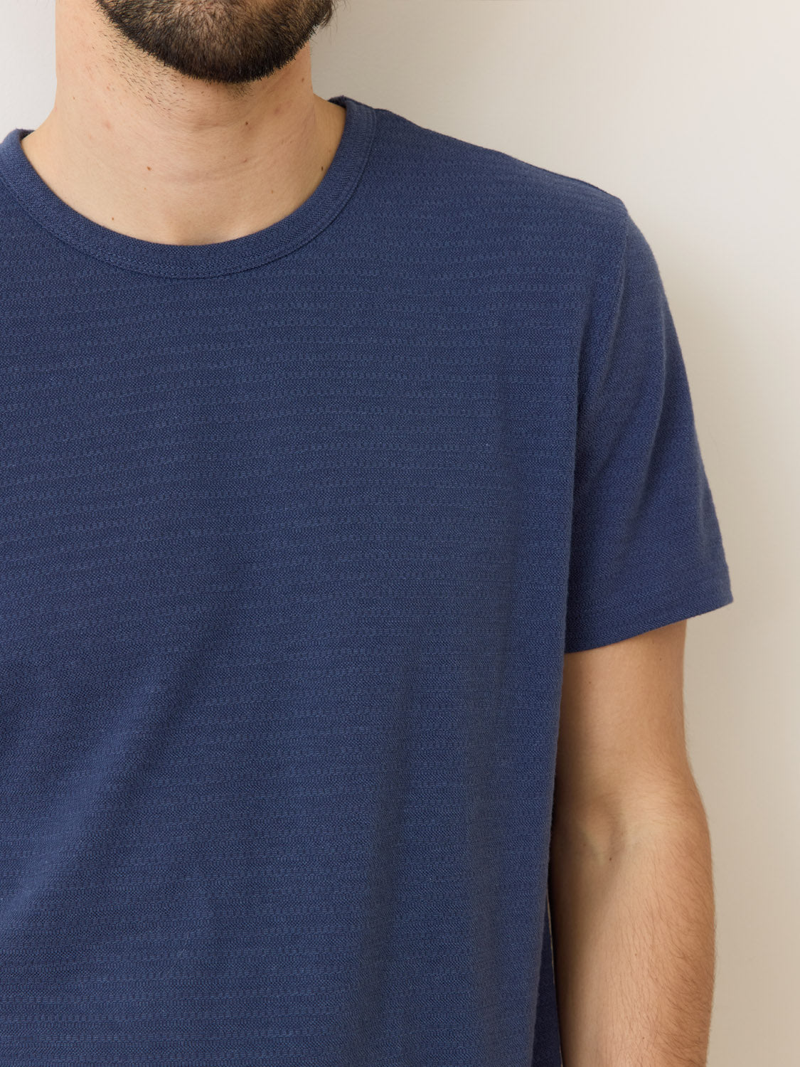 A person is partially visible against a plain background, wearing a Marine Layer Signature Jacquard Crew Tee in navy blue made from recycled polyester. Only the right side of their upper body and part of their bearded face are shown.