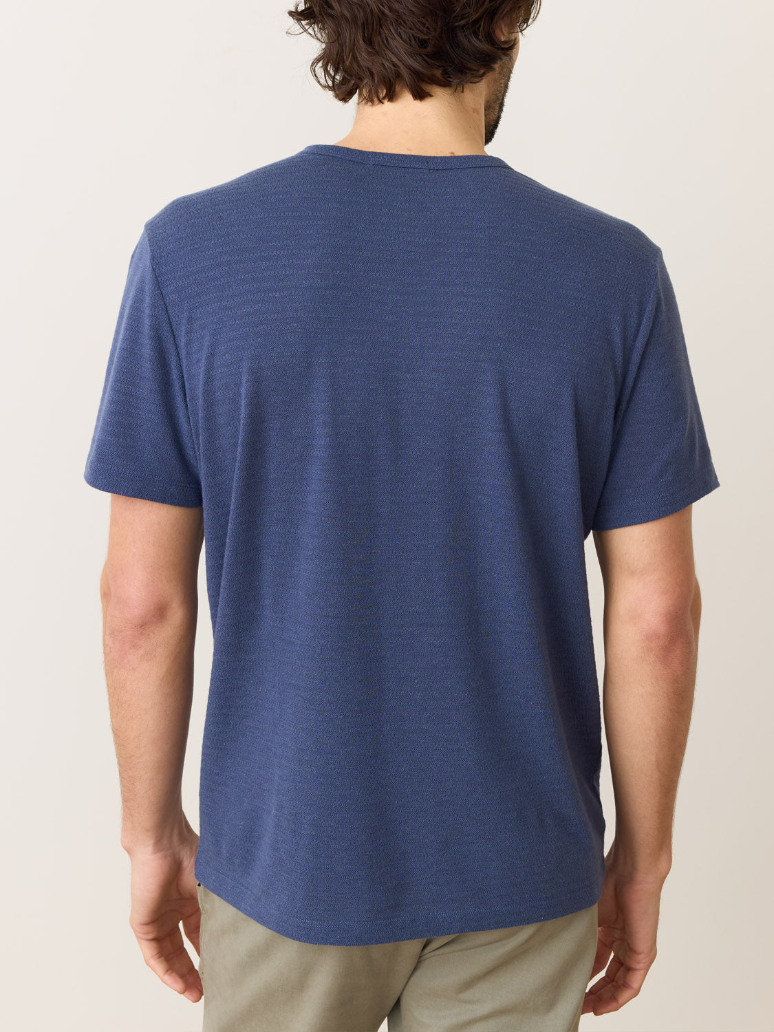 Someone with dark hair wears a Marine Layer Signature Jacquard Crew Tee in blue and beige pants, facing away from the camera against an off-white background.
