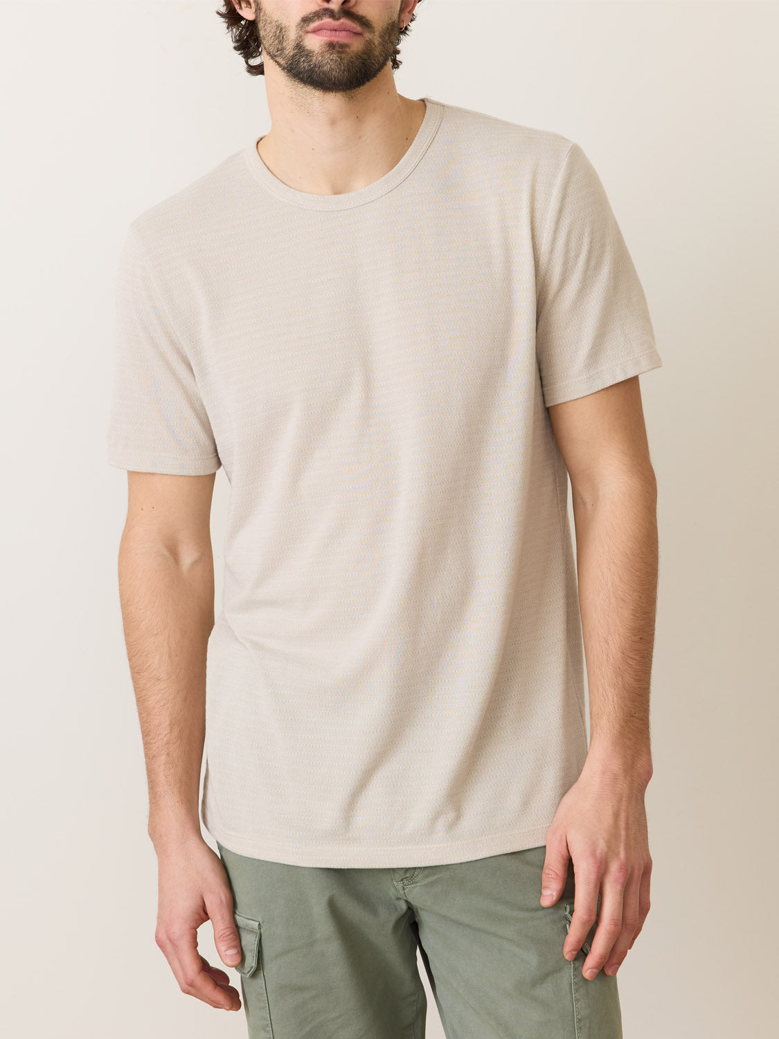 A person wearing a Marine Layer Signature Jacquard Crew Tee in beige, made from recycled polyester, along with olive green pants, stands against a neutral background. The individuals face is partially out of frame.
