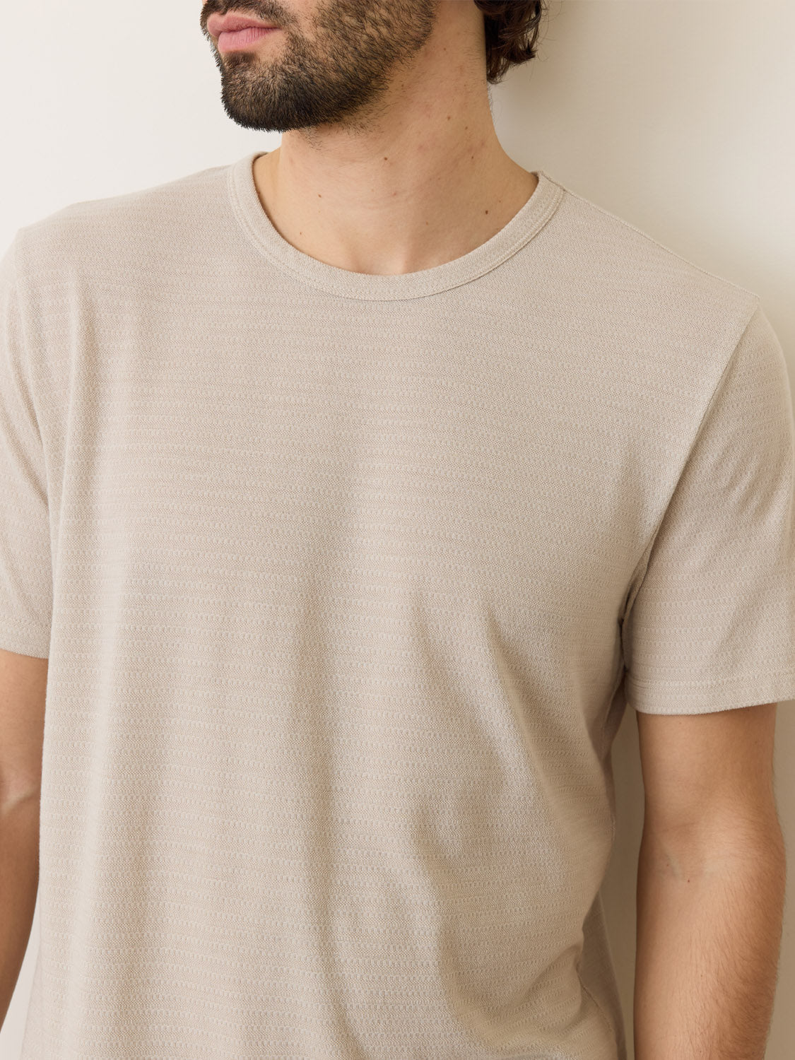A man in a light beige Marine Layer Signature Jacquard Crew Tee is facing partially away against a plain background. His face is not fully visible, and hes positioned slightly off-center, highlighting the shirts intricate fabric pattern.