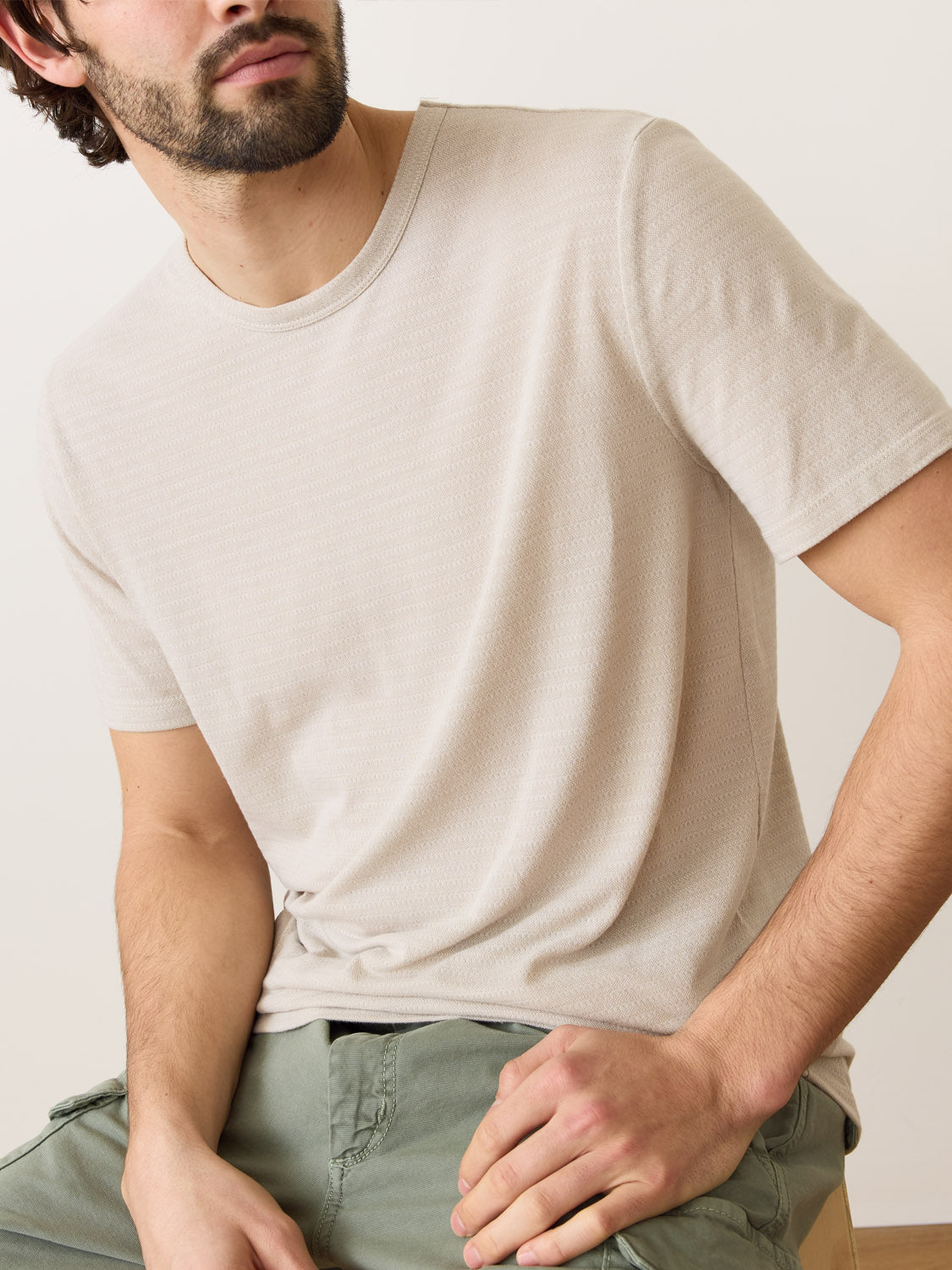 A person is sitting, wearing a Marine Layer Signature Jacquard Crew Tee and light green pants. The photo shows them from the shoulders down against a plain background, with arms resting casually to suggest relaxation and comfort.