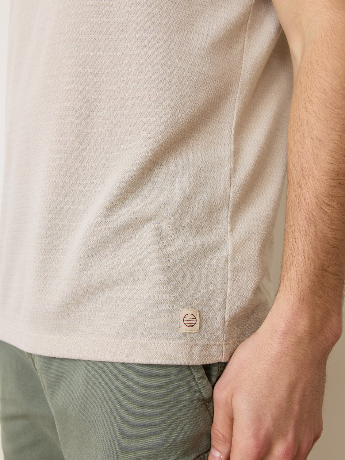 Close-up of someone wearing the Marine Layer Signature Jacquard Crew Tee in beige with olive green pants. The tee, made from recycled polyester, has a small circular logo on the lower side, and the persons arm is visible on the right side of the image.