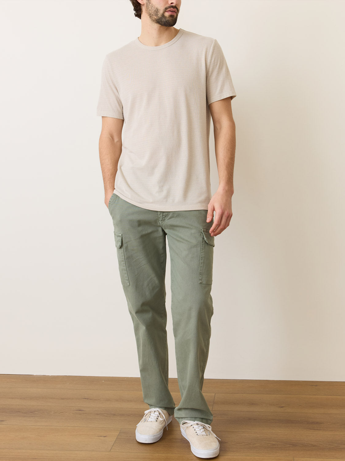 A person wears a light beige Marine Layer Signature Jacquard Crew Tee crafted from recycled polyester with olive cargo pants and white sneakers, standing on a wooden floor against an off-white wall, glancing to the side.