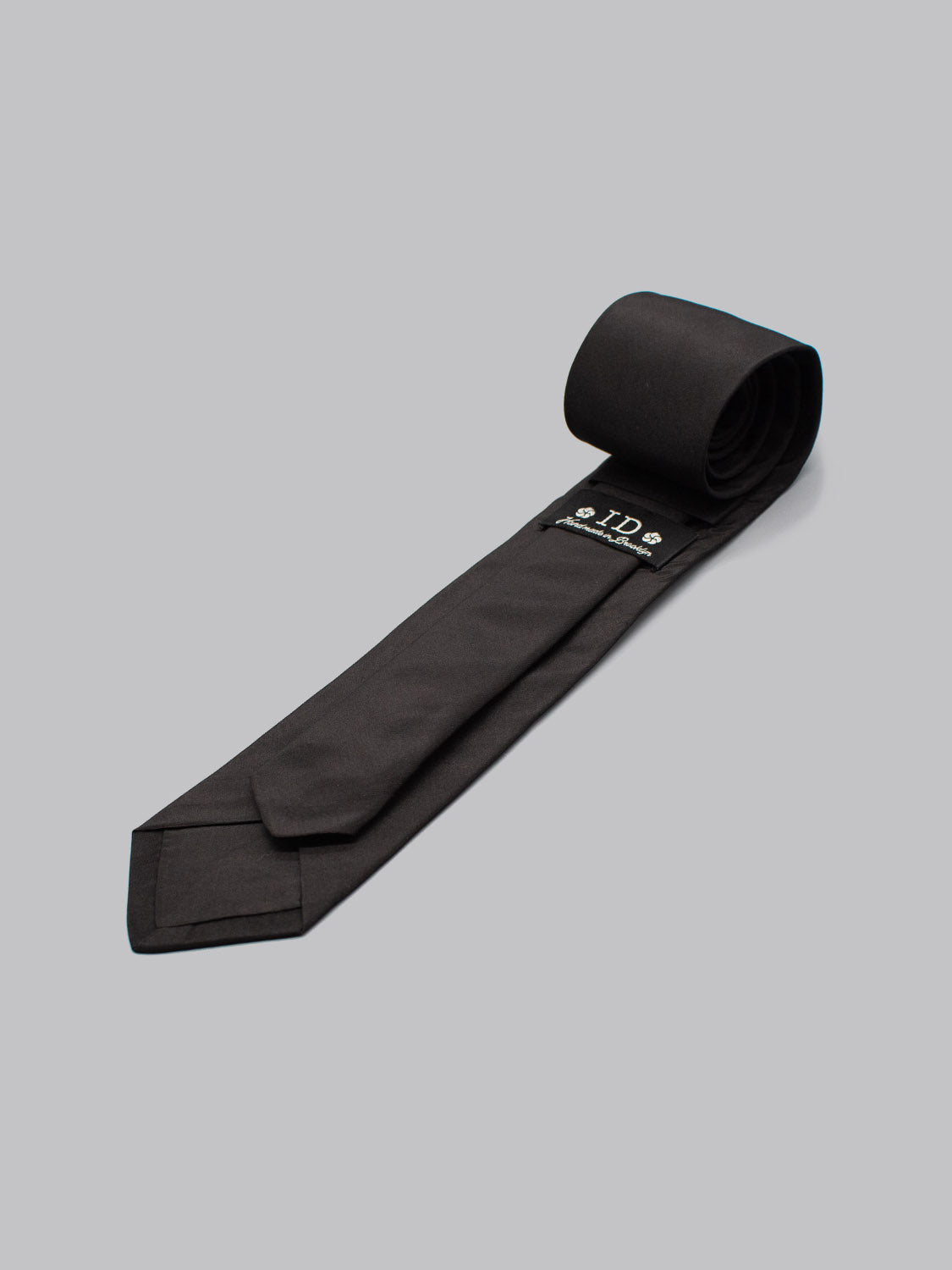 The ID Menswear ID Silk Taffeta Tie in black is rolled on one end and set against a light grey background, showing its branded label sewn on the back—ideal for formal occasions.