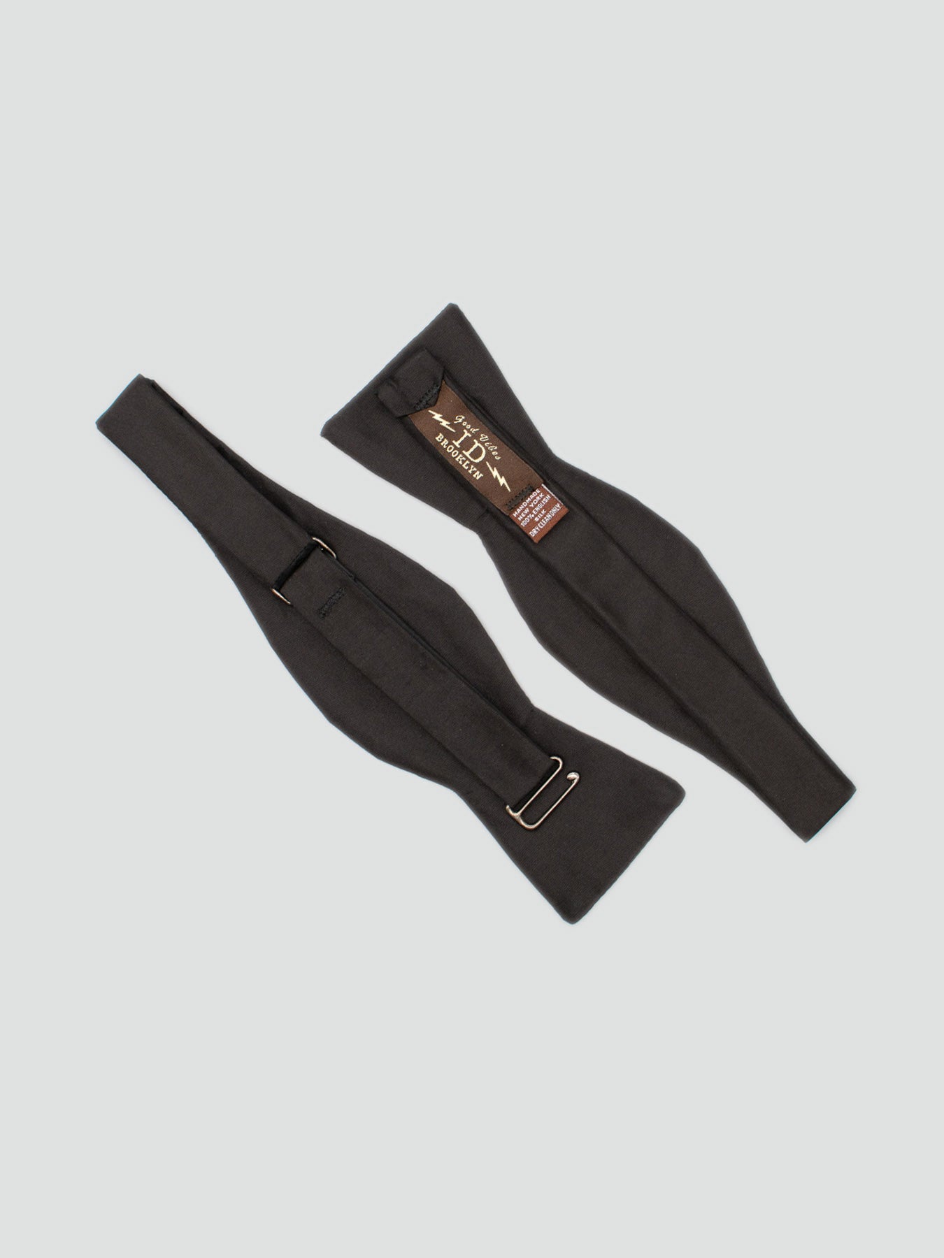 Two black Silk Taffeta Bow Ties by ID Menswear, handmade in Brooklyn with a slightly padded look, are laid flat on a light grey background. One displays the label and metal clip; the other shows the reverse side.