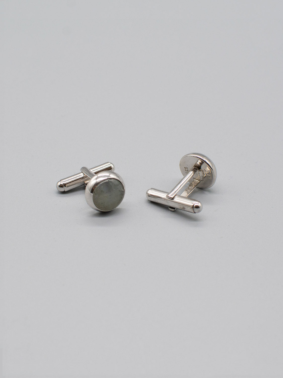 Two Silver Gemstone Cufflinks from ID Menswear rest on a light gray surface, each showcasing a round, flat face with a reflective finish and rhodium plating, attached to a T-bar closure. The minimalistic design exudes modern elegance.