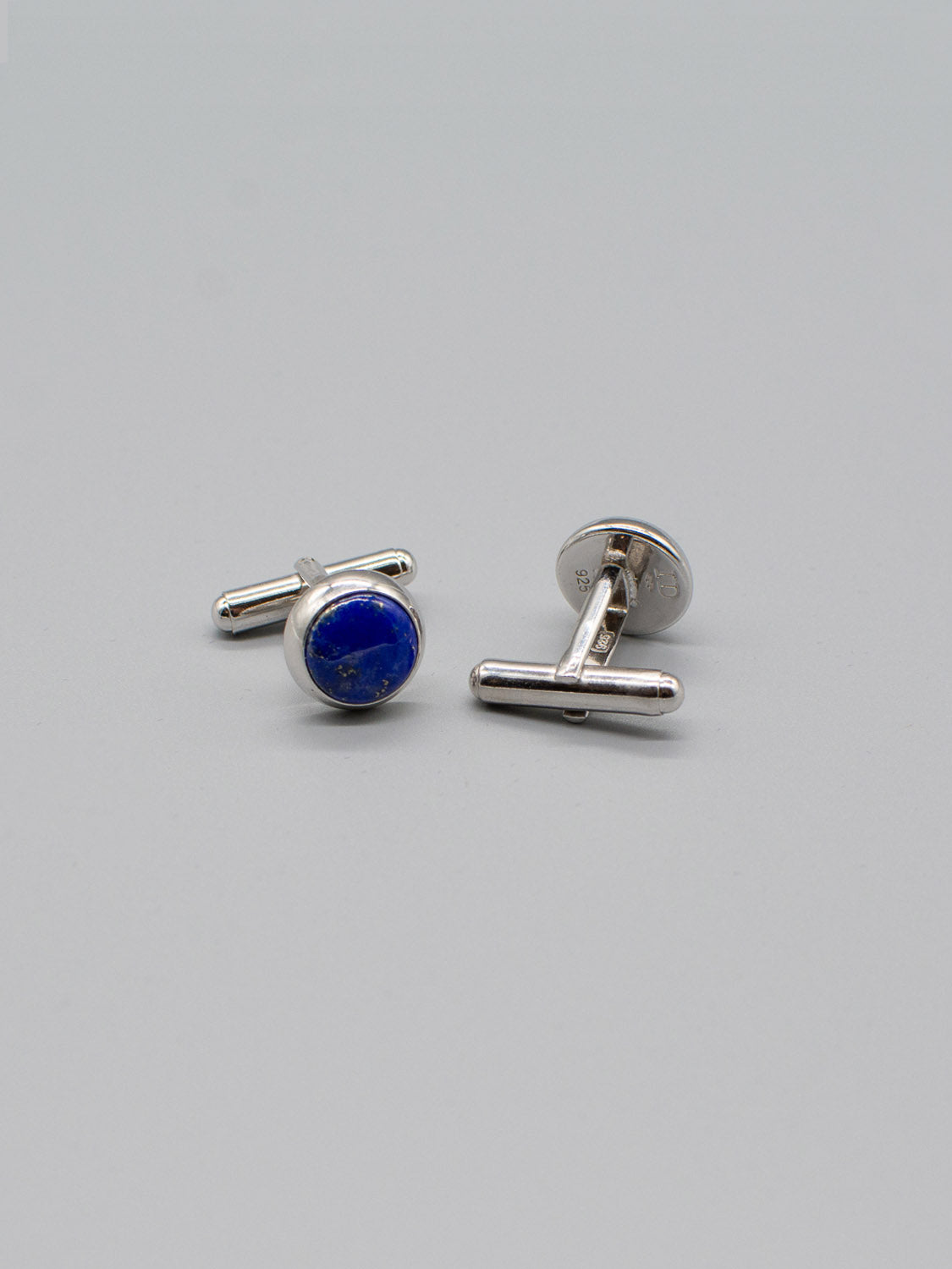Two Silver Gemstone Cufflinks by ID Menswear with round blue stones adorn a plain gray background. One cufflink stands upright, the other reclines, highlighting its captivating gemstone inlay.