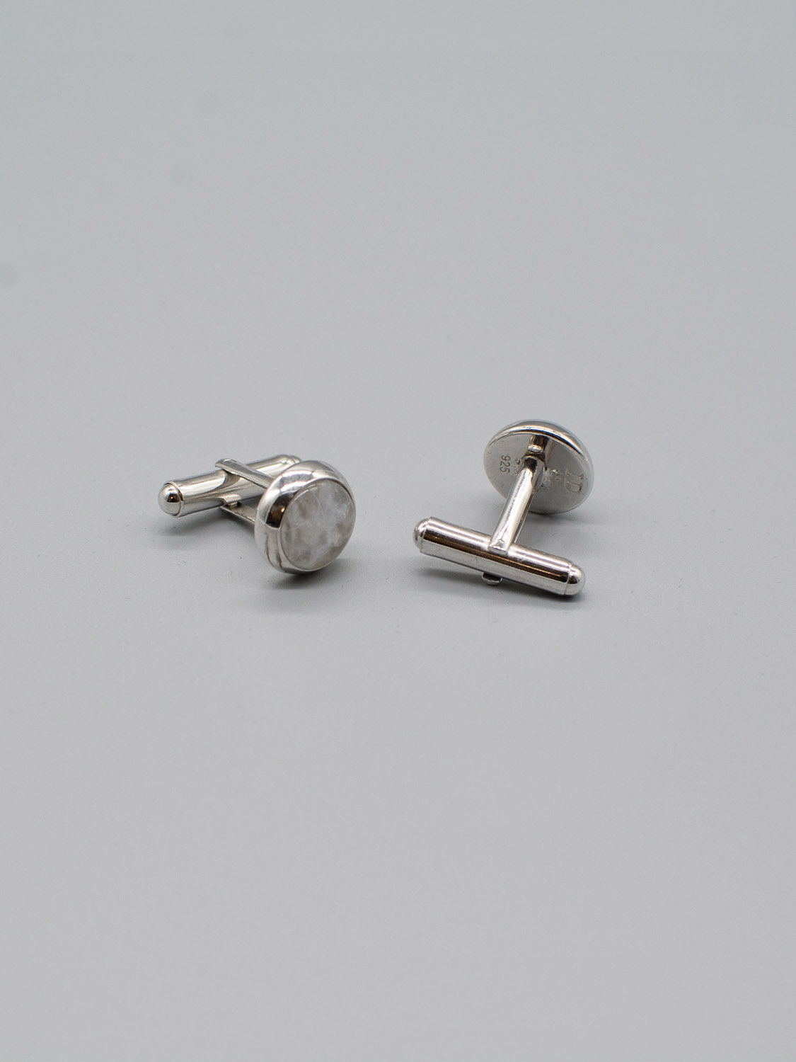 A pair of Silver Gemstone Cufflinks from ID Menswear, featuring round, marble-like tops with subtle gemstone inlay, rest on a smooth light gray surface. One cufflink is upright showing its toggle, while the other lies flat displaying its polished front.