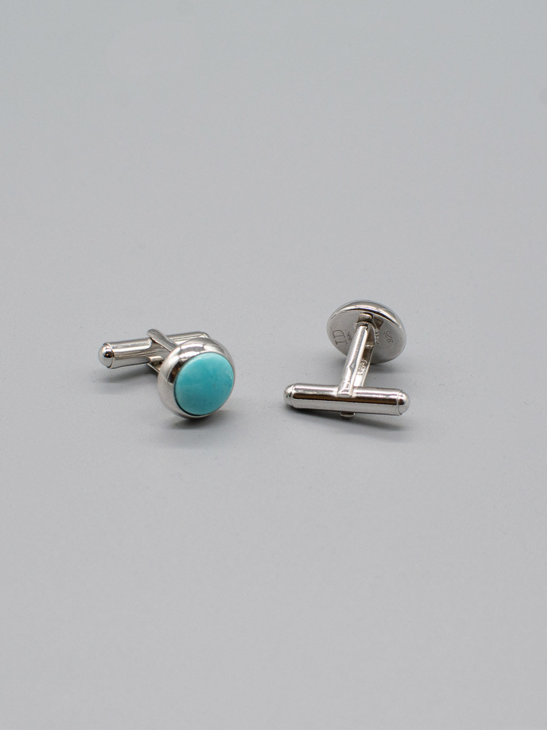 Two Rhodium Plated Silver Gemstone Cufflinks by ID Menswear are set against a plain gray background. One stands upright, highlighting the circular turquoise stone, while the other lies on its side to reveal the T-shaped backing.