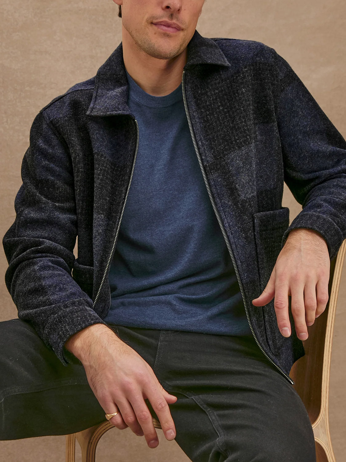 A man sits on a wooden chair wearing a Rails Simon Jacket—a dark wool blend zip-up—over a blue T-shirt and black pants. His face is partially out of frame, with his right hand on his knee against a beige background.