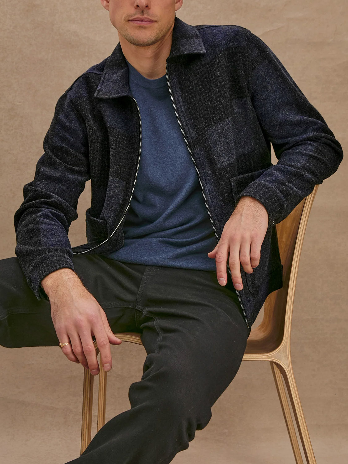 A man sits on a wooden chair against a neutral background, wearing the Rails Simon Jacket over a blue shirt and black pants, with one arm resting on the chair and the other on his knee.