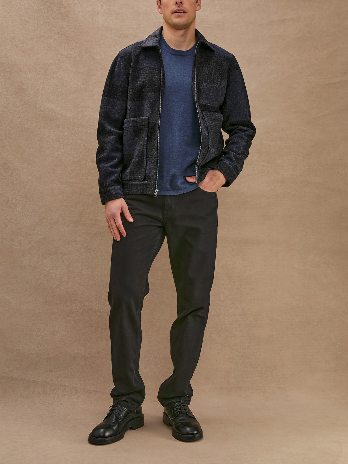 A man stands against a beige backdrop wearing the Rails Simon Jacket, a navy-blue wool blend zip-up, paired with a blue t-shirt, black pants, and black shoes. His left hand is in his pocket and his face is partially out of frame.