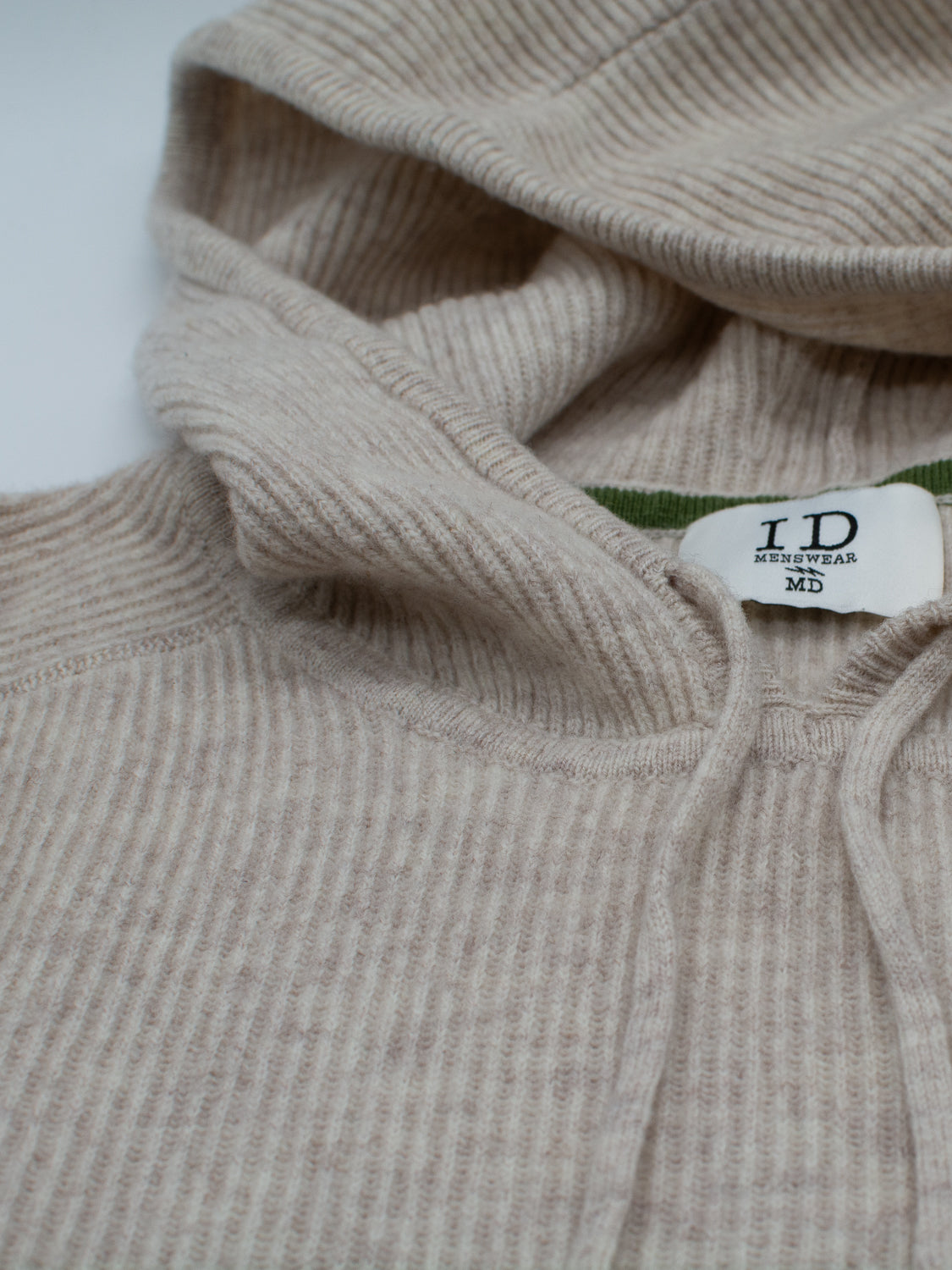 A close-up of the Skien Merino Wool Hoody by ID Menswear—a beige half-cardigan knit with hood and drawstring, ideal for cold weather. Inside, a white tag reads ID Menswear MD beside a green inner trim.