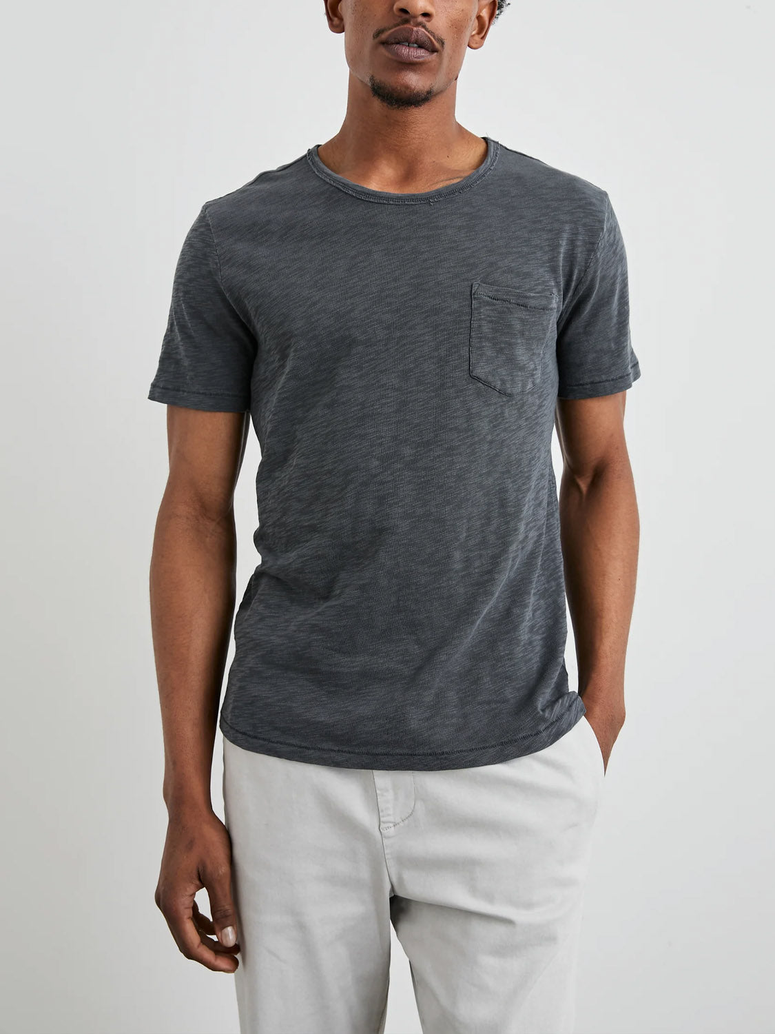 Someone wears a Rails Skipper Tee in dark gray, featuring a vintage wash, super soft cotton, and a chest pocket, paired with light gray pants. They stand against a plain white background, highlighting its versatile layering potential.