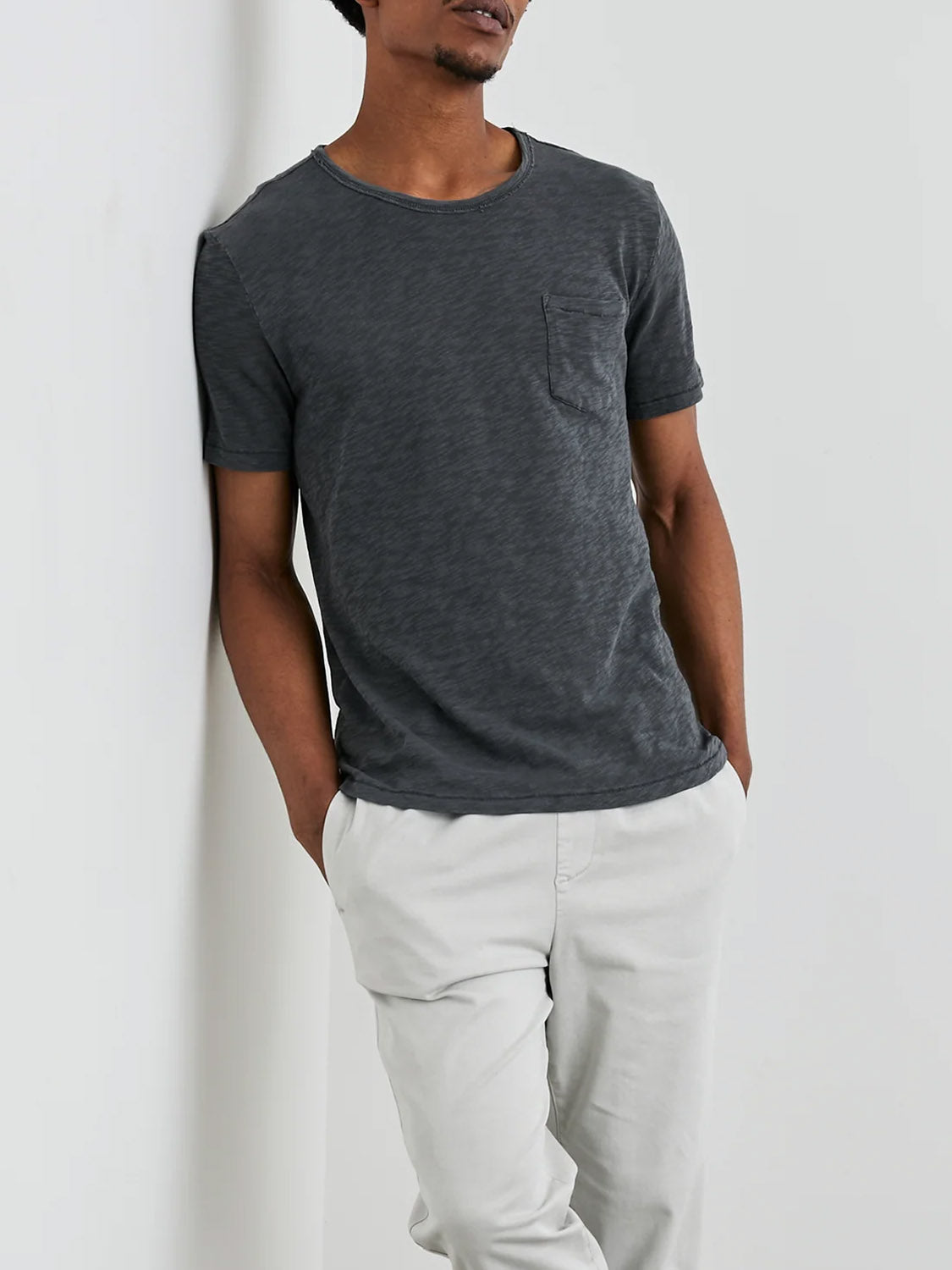 A person stands against a white wall, wearing the Rails Skipper Tee in dark gray with a vintage wash and light gray pants. This super soft cotton tee features a chest pocket. With hands tucked in their pockets, they exude a relaxed and effortlessly stylish vibe.