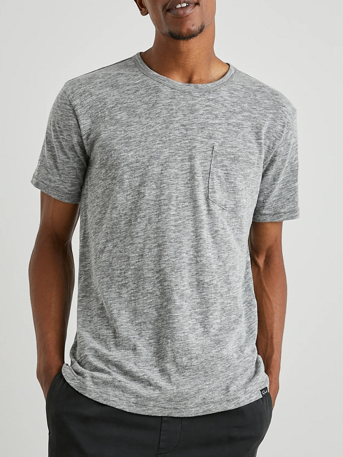A person is wearing the Rails Skipper Tee, a gray short-sleeved t-shirt crafted from super soft cotton with a chest patch pocket, paired with black pants. They stand against a plain background with hands in pockets.