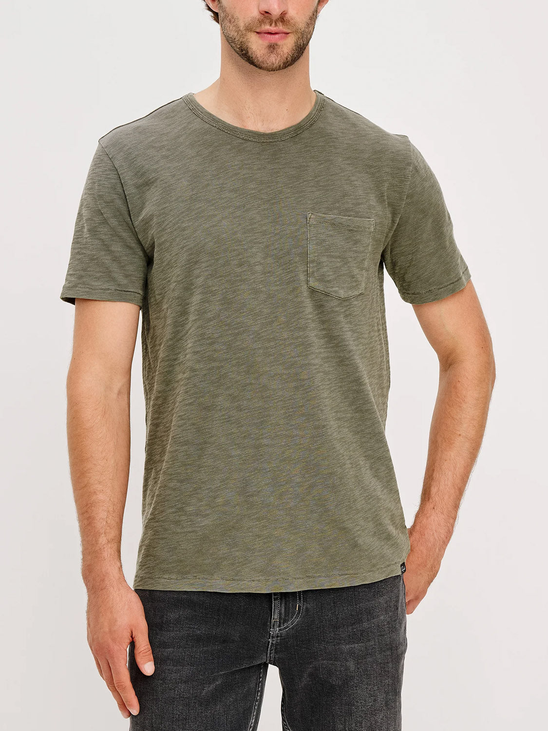 A man with a trimmed beard stands against a plain background wearing the Rails Skipper T-Shirt in olive green, featuring a chest pocket, paired with dark jeans. His face is partially visible.