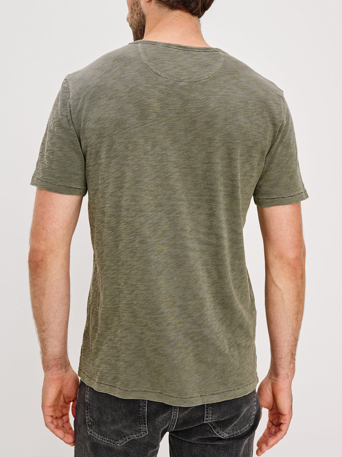A man stands with his back to the camera, wearing the Rails Skipper T-Shirt in olive green and dark gray jeans, set against a plain white background.