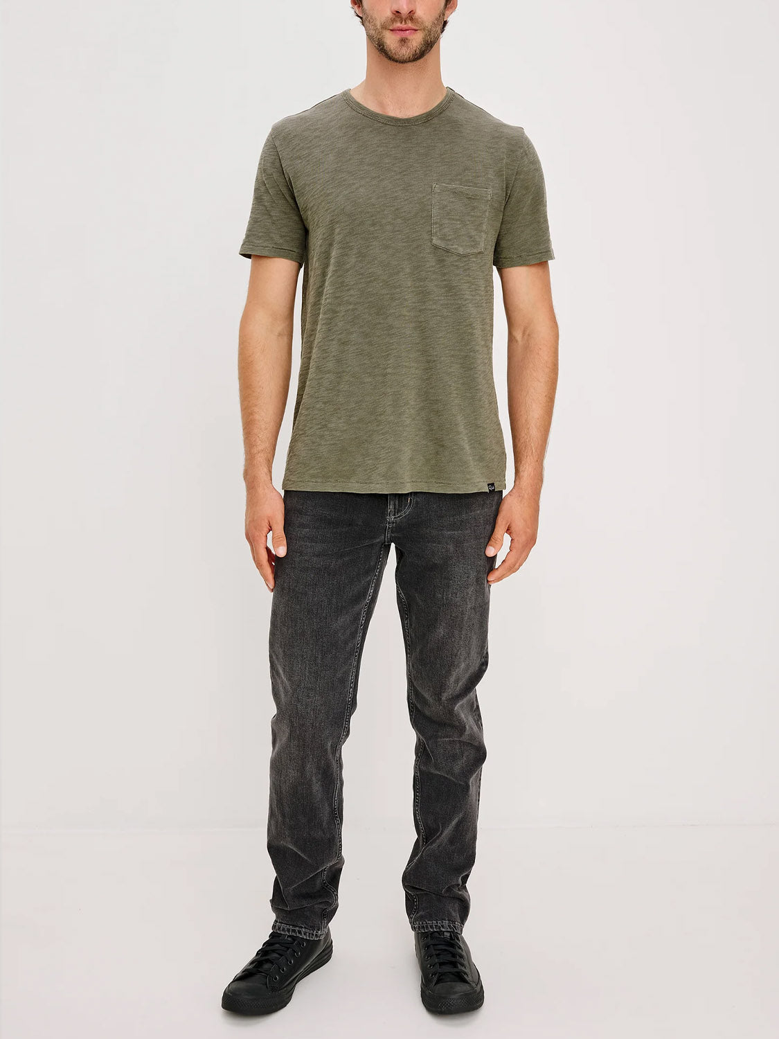 A man stands against a plain white background, wearing the Rails Skipper T-Shirt—a super soft cotton green short-sleeve tee with a chest pocket—paired with dark grey jeans and black sneakers. His arms are relaxed by his sides.