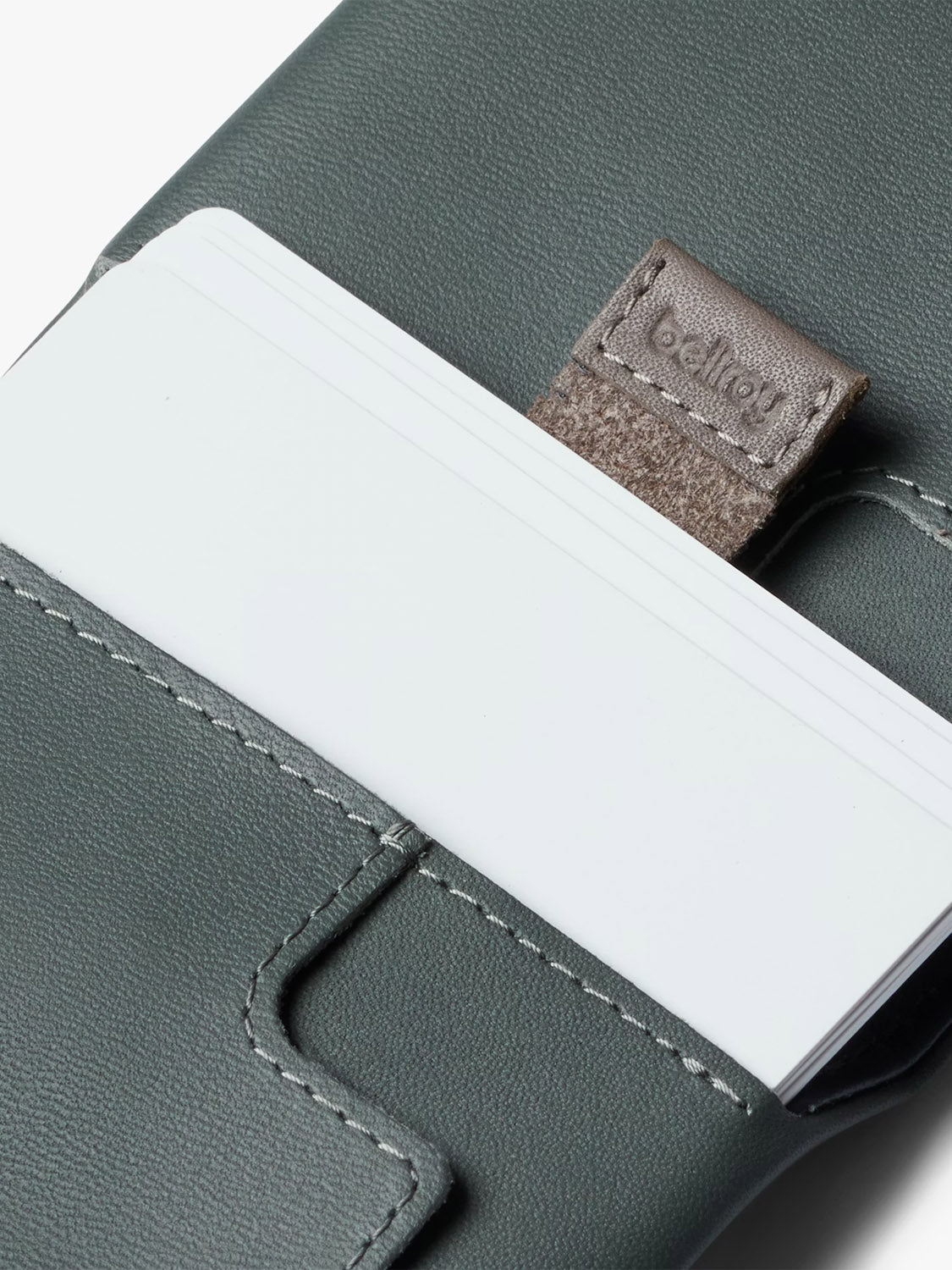 A close-up of white cards partially inserted into Bellroy's Slim Sleeve Wallet, a slim leather card holder with white stitching and a brown tab.