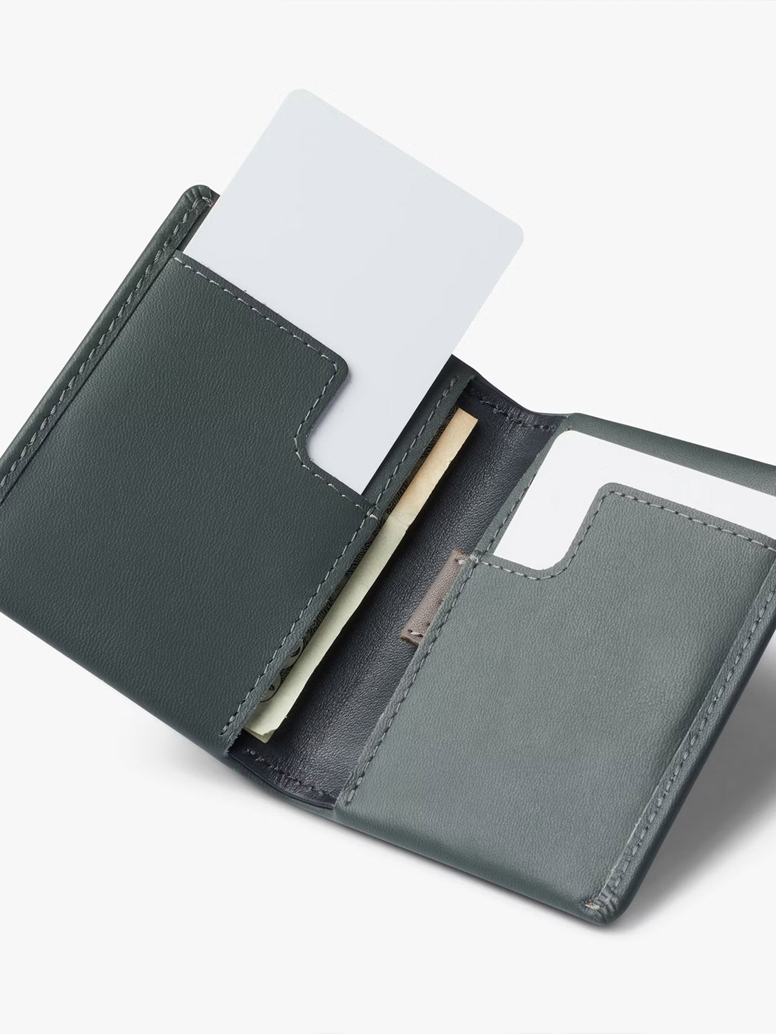 The Bellroy Slim Sleeve Wallet in minimalist dark green leather lies open on a plain light background, showing two pockets—one with a white card, the other with a white card and folded cash.