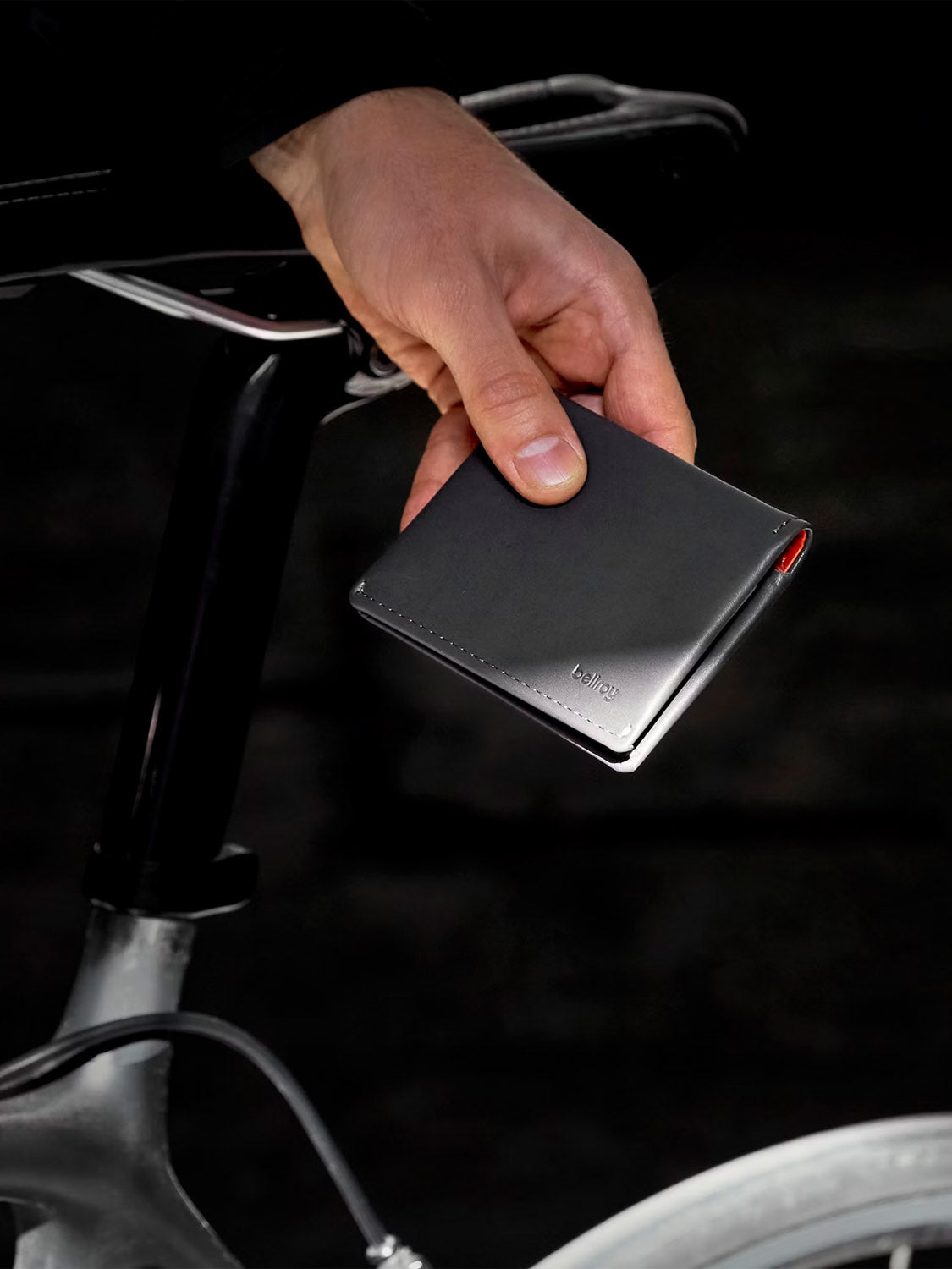 A person holds a closed Bellroy Slim Sleeve Wallet beside a bicycle seat, their hand resting on the saddle rail. The dark background accentuates both the wallet and the bike seat.