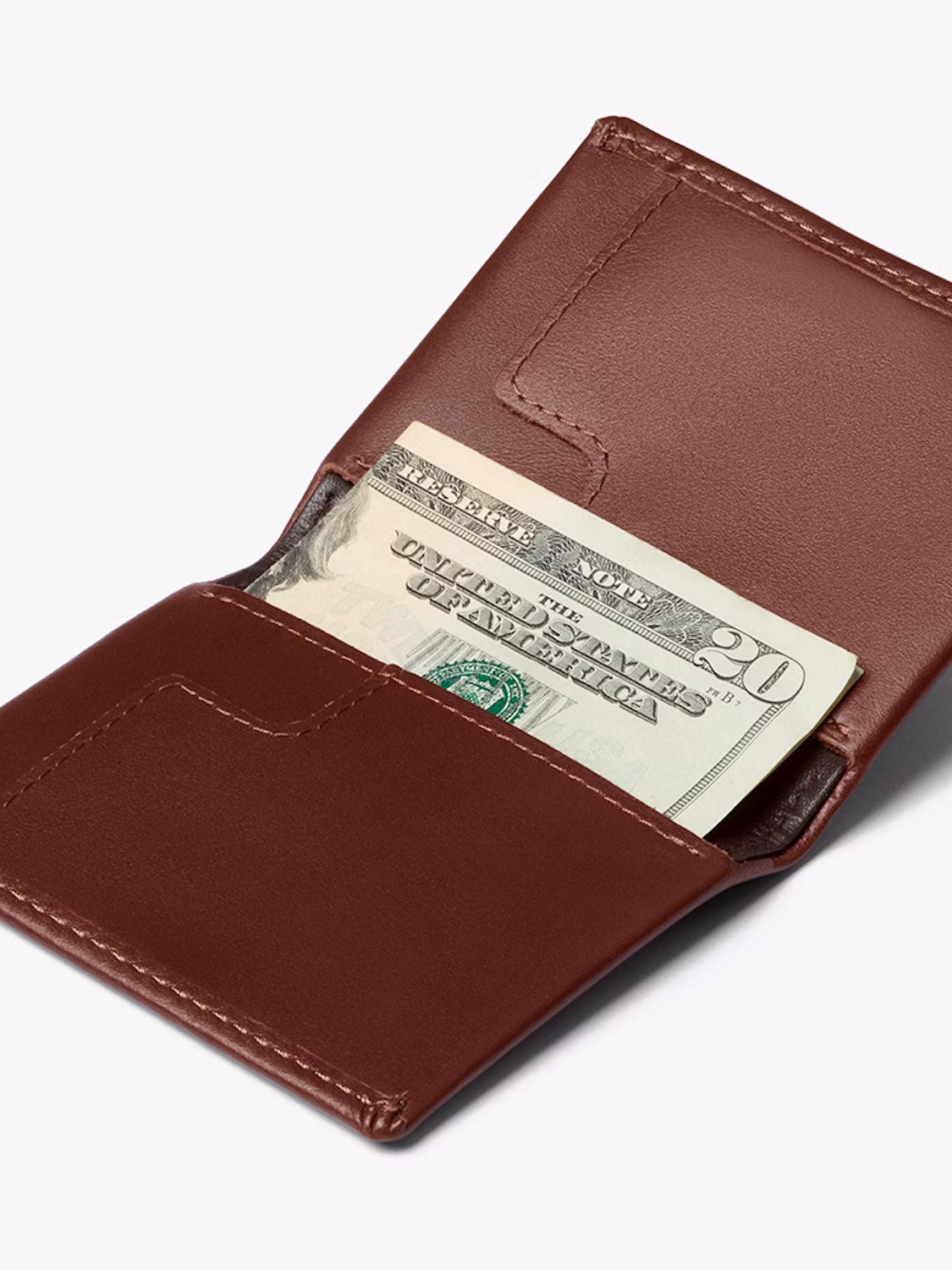 A Bellroy Slim Sleeve Wallet in brown sits open against a light background, displaying a visible twenty-dollar bill inside.