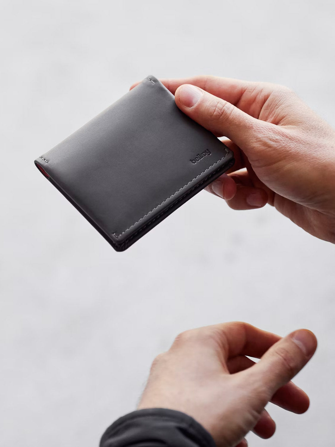 A person holds a closed Bellroy Slim Sleeve Wallet with both hands against a softly blurred, light background.