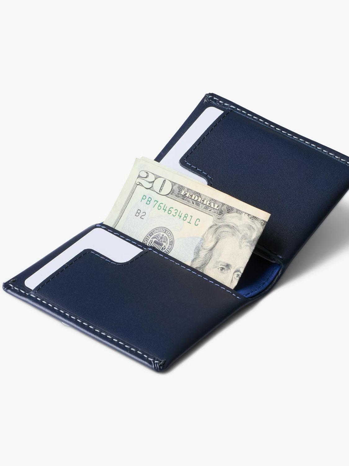The Bellroy Slim Sleeve Wallet in navy blue with white stitching is open on a plain white background, displaying a twenty dollar bill and two white cards inside.
