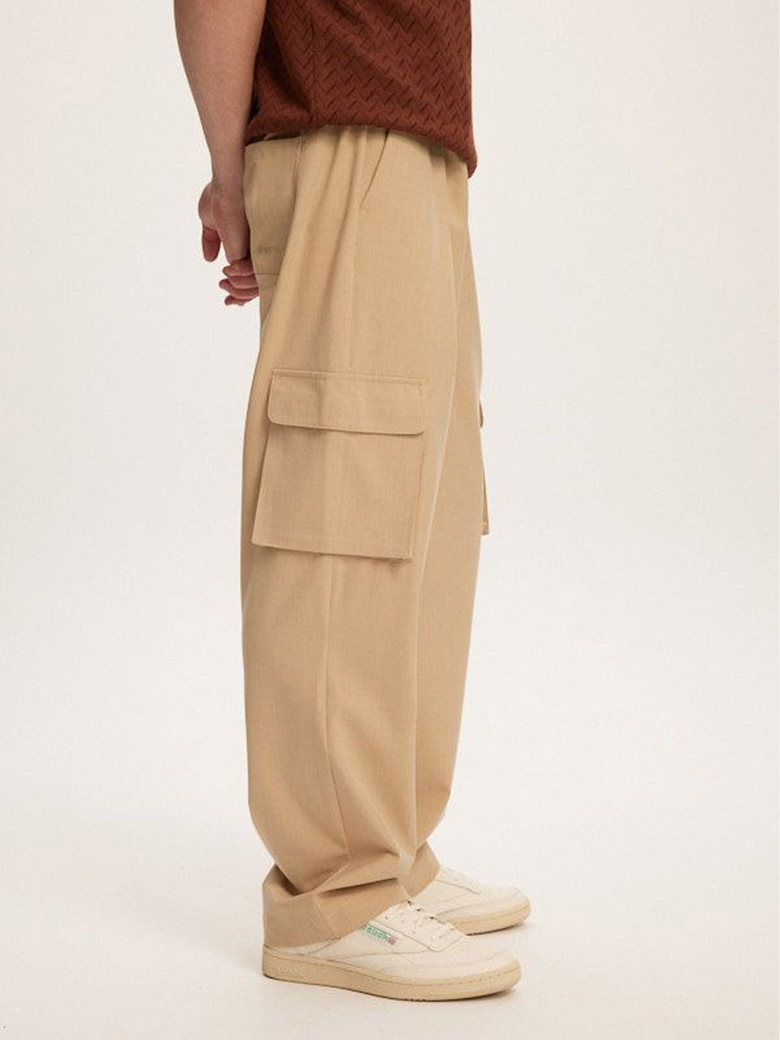A person stands sideways on a plain background, wearing Kuwalla Smart Cargo Pants in beige with large pockets and white sneakers, perfectly complementing a tailored brown patterned shirt.