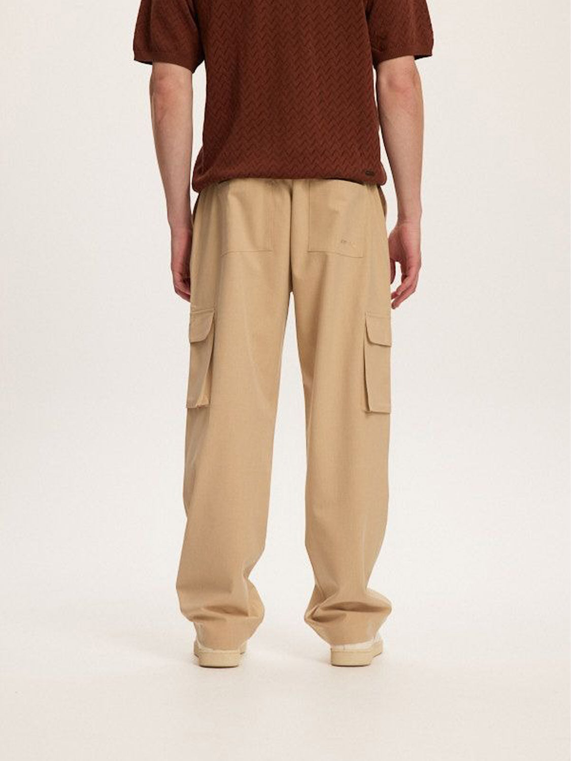 A person faces away from the camera, highlighting their style in a brown short-sleeve top and Kuwalla Smart Cargo Pants by Kuwalla in beige, featuring practical side pockets. The background is plain off-white.