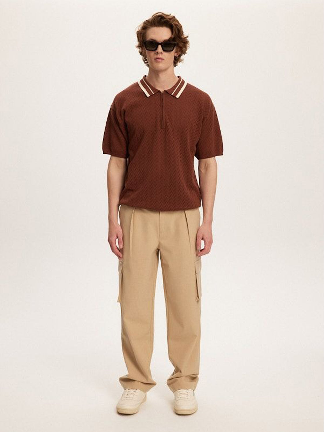 A person with shoulder-length brown hair stands against a plain background wearing sunglasses, a brown short-sleeve polo shirt with a white-striped collar, light-colored sneakers, and versatile Kuwalla Smart Cargo Pants in beige.
