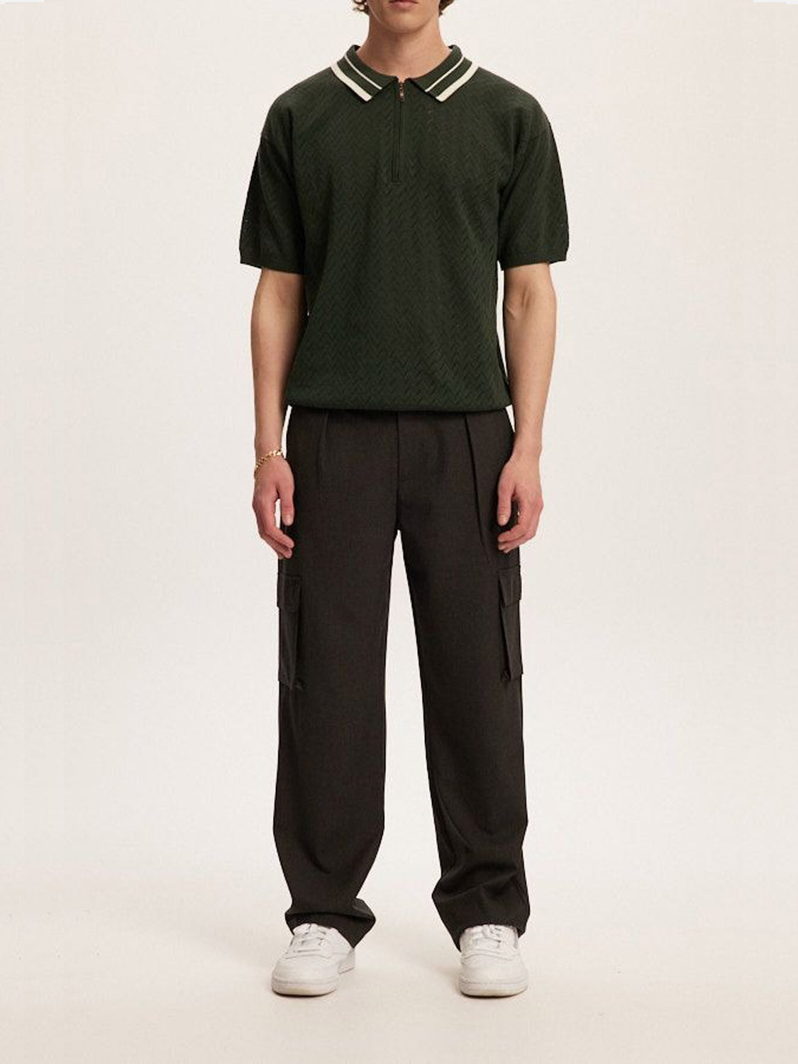 Wearing a dark green short-sleeved polo shirt with a white-striped collar and tailored fit, a person pairs it with Kuwalla Smart Cargo Pants from Kuwalla and white sneakers. They stand against a plain light background, showcasing versatile style.