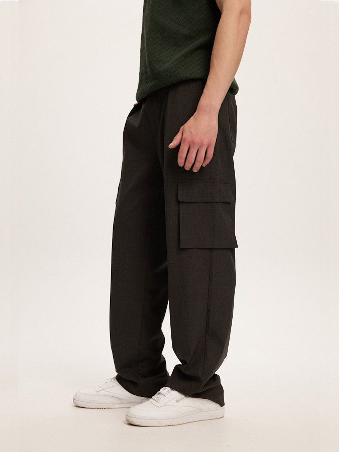 Wearing a dark green textured top, white sneakers, and Kuwalla Smart Cargo Pants in black with a versatile design and large side pockets, the individual stands against a plain backdrop. They effortlessly blend function with style.