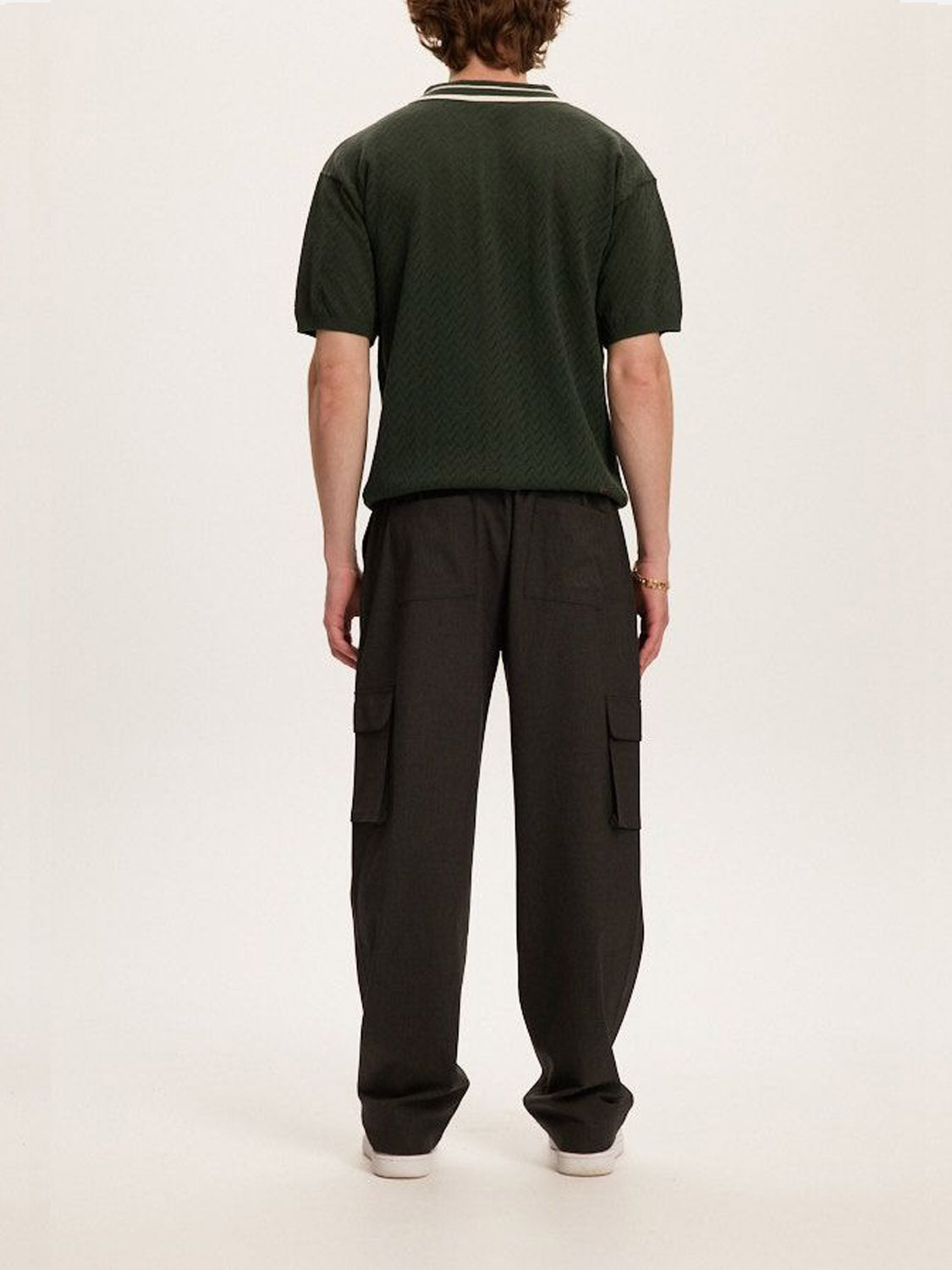 A person stands facing away, highlighting a dark green tailored short-sleeve shirt paired with the versatile Kuwalla Smart Cargo Pants from Kuwalla. Their curly hair complements the style, and sneakers ground them against a plain white background.