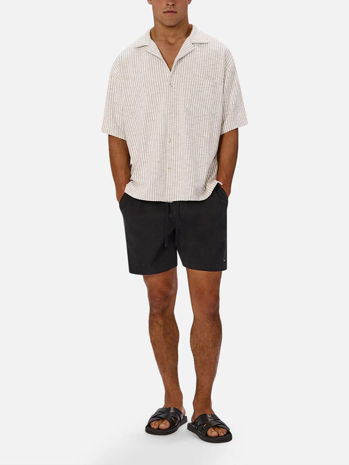 A man wears the Industrie Sonora Short Sleeve Shirt in beige, paired with black shorts and sandals, standing with hands in pockets against a plain white background.