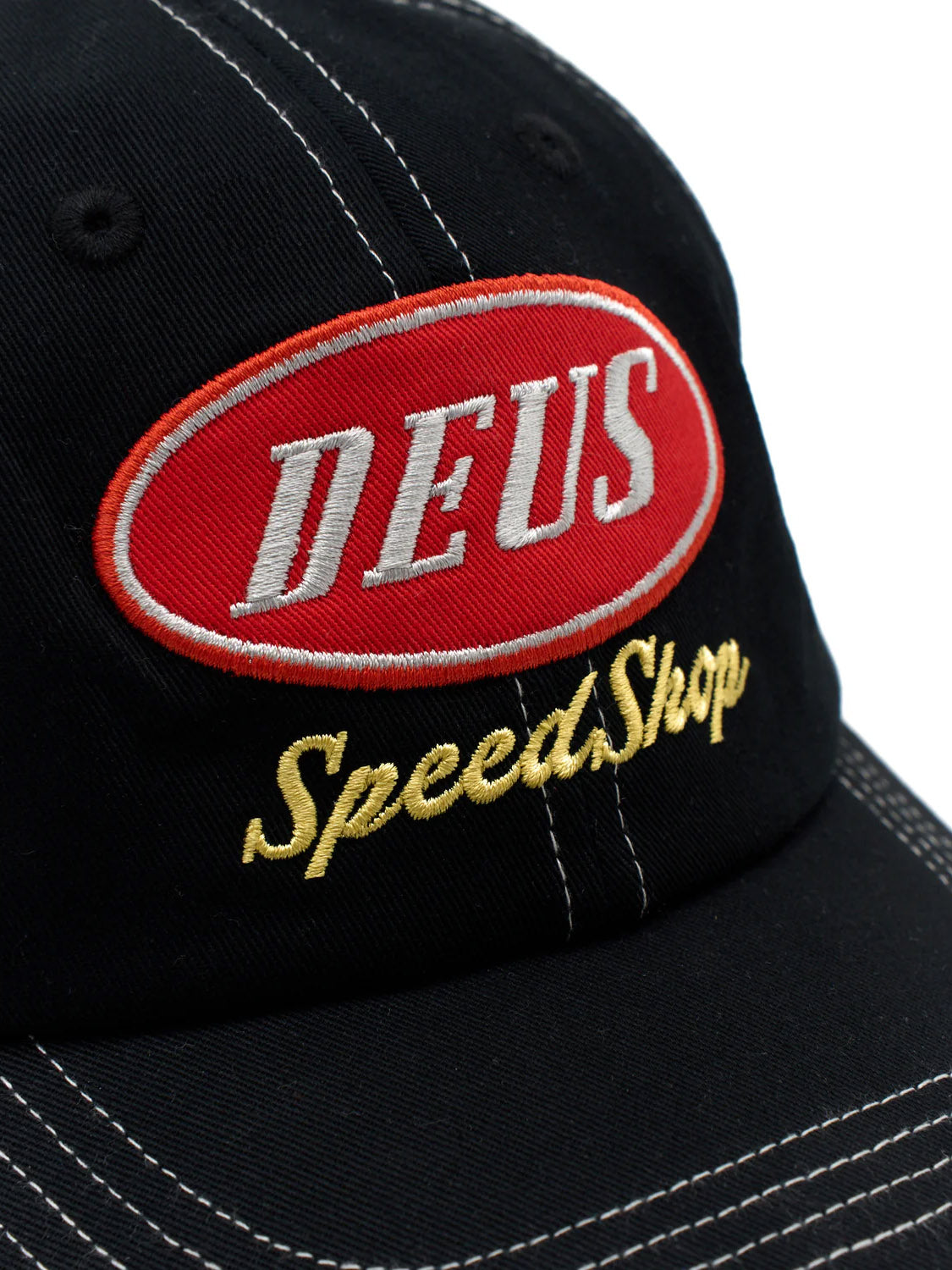 The Deus Speedshop Cap is a black cotton twill dad cap with white stitching, featuring a red oval patch with "DEUS" embroidered in white and "Speed Shop" stitched below in yellow.