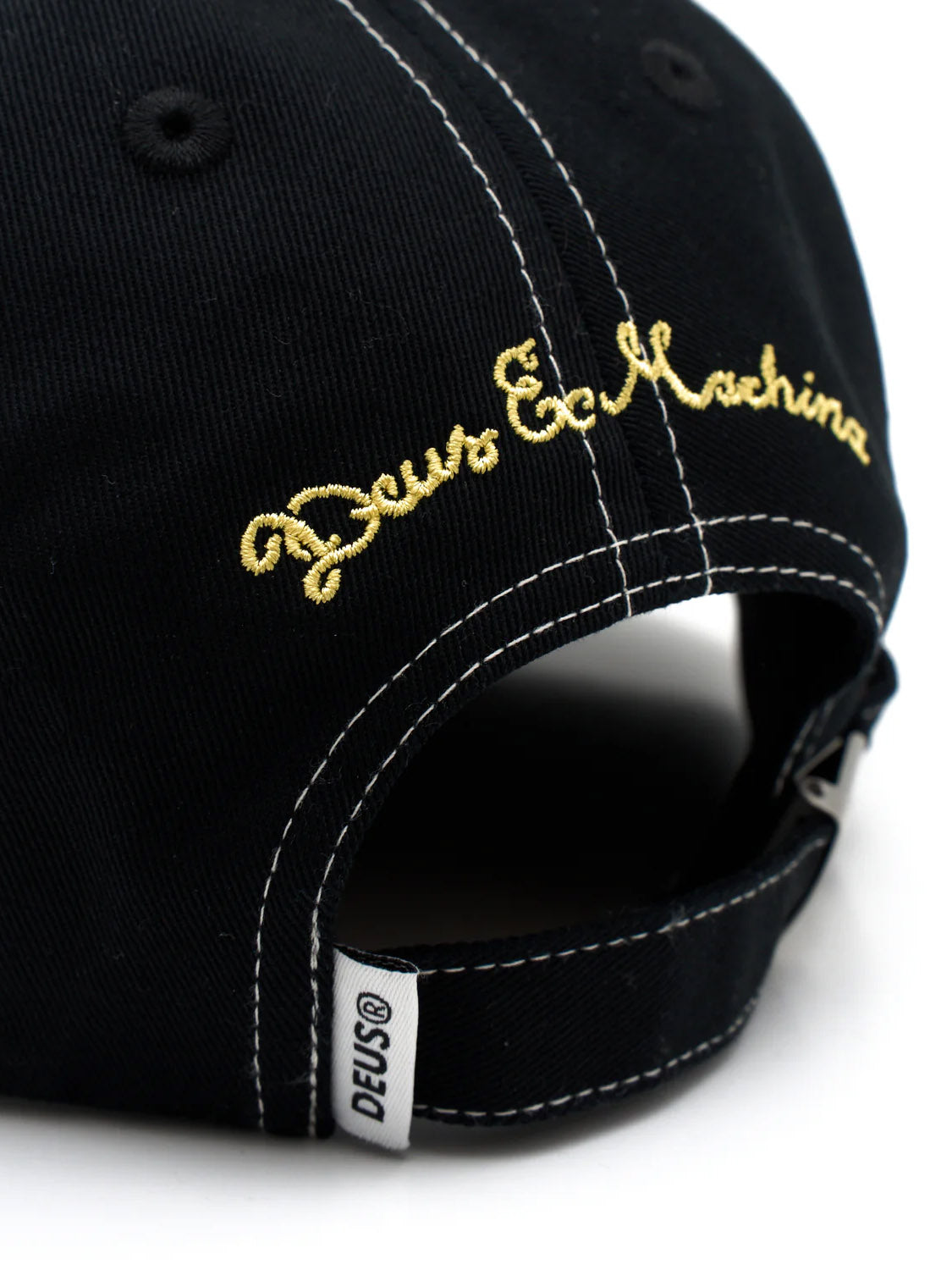 Close-up of the back of a black cotton twill Speedshop Cap by Deus, featuring yellow "Deus Ex Machina" embroidery and a DEUS® fabric tag near the adjustable strap.