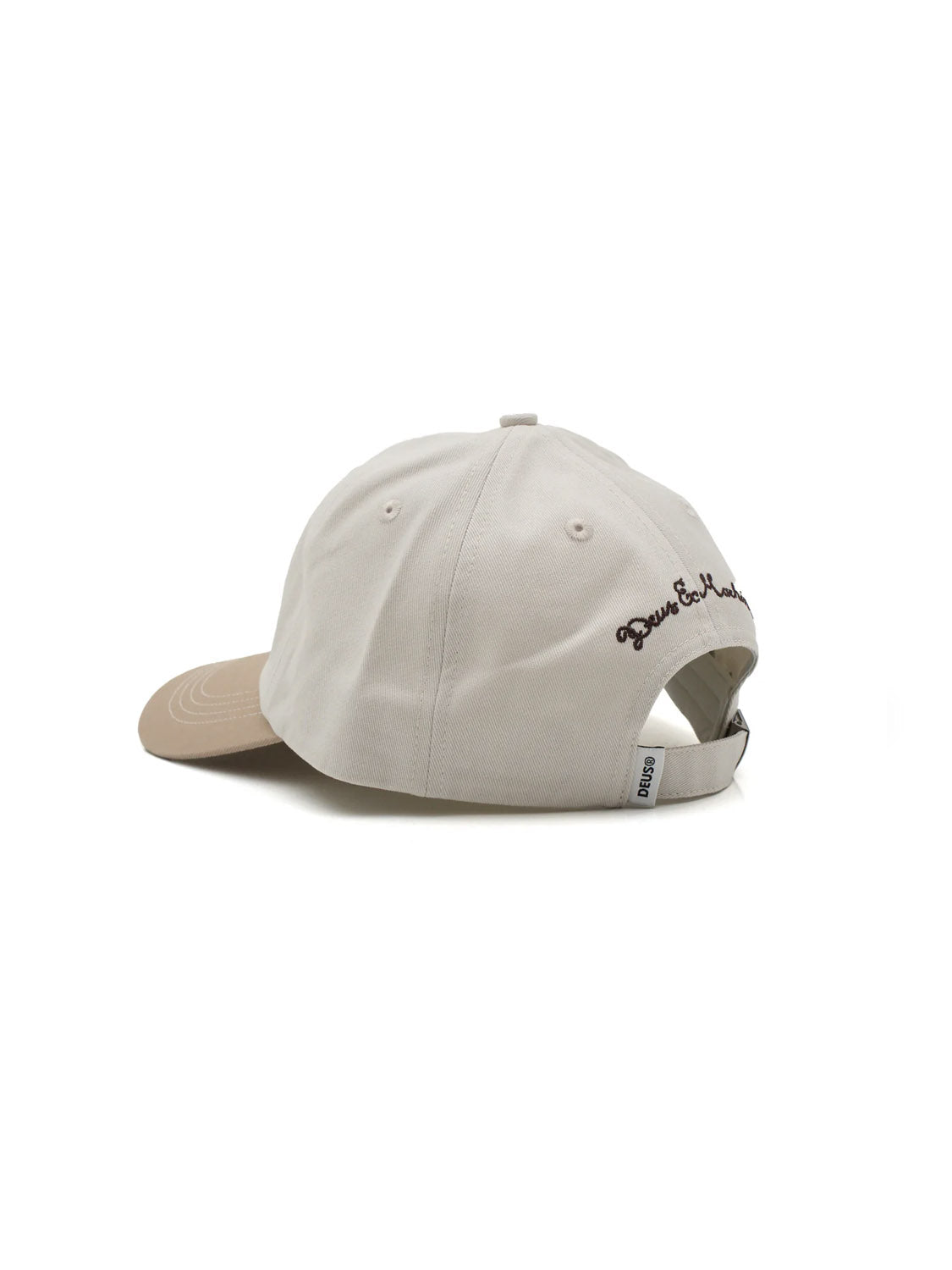 The Deus Speedshop Cap is a beige and light tan cotton twill dad cap with an adjustable strapback, black cursive embroidery above the opening, and a small white tag, shown from a rear-side angle.