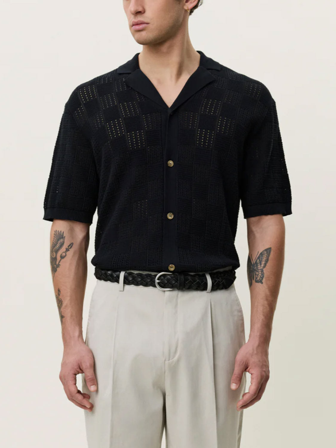 A person wears the Les Deux Squared Knitted SS Shirt in black, paired with light gray pants and a black braided belt. Their tattooed arms stand out against a plain off-white background.