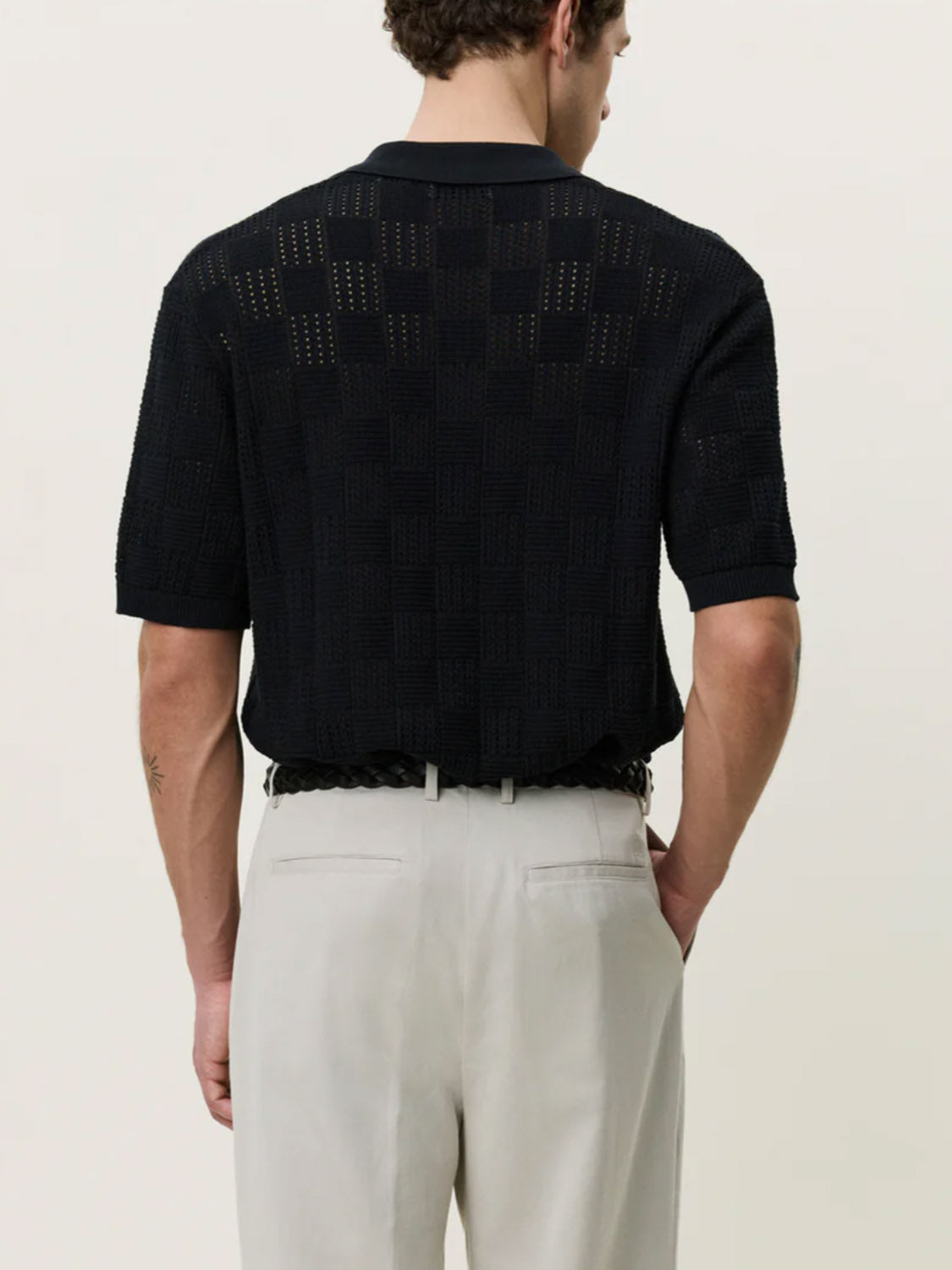 A man is seen from behind wearing the Les Deux Squared Knitted SS Shirt—a black, short-sleeve cotton top with subtle checks and perforations—paired with light beige trousers and a braided black belt.